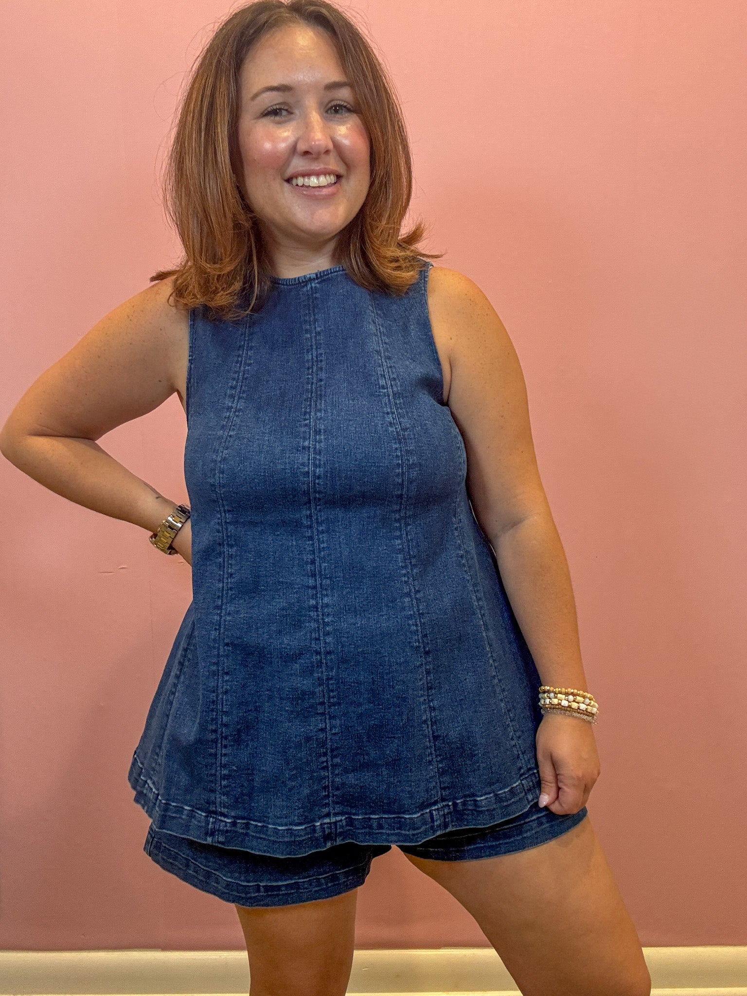 A woman modeling a sleeveless blue denim romper with matching denim shorts, standing against a solid pink wall.