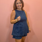 A woman modeling a sleeveless blue denim romper with matching shorts, standing against a solid pink wall.