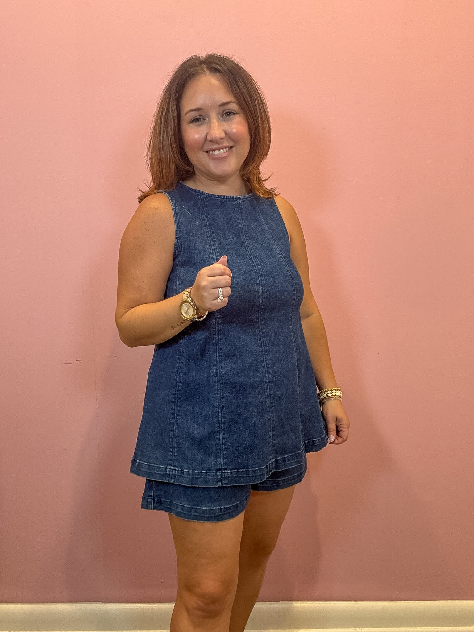 A woman modeling a sleeveless blue denim romper with matching shorts, standing against a solid pink wall.