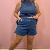 A woman modeling a matching two-piece denim set consisting of a sleeveless high-neck top and elastic-waist shorts, posed against a solid pink wall.