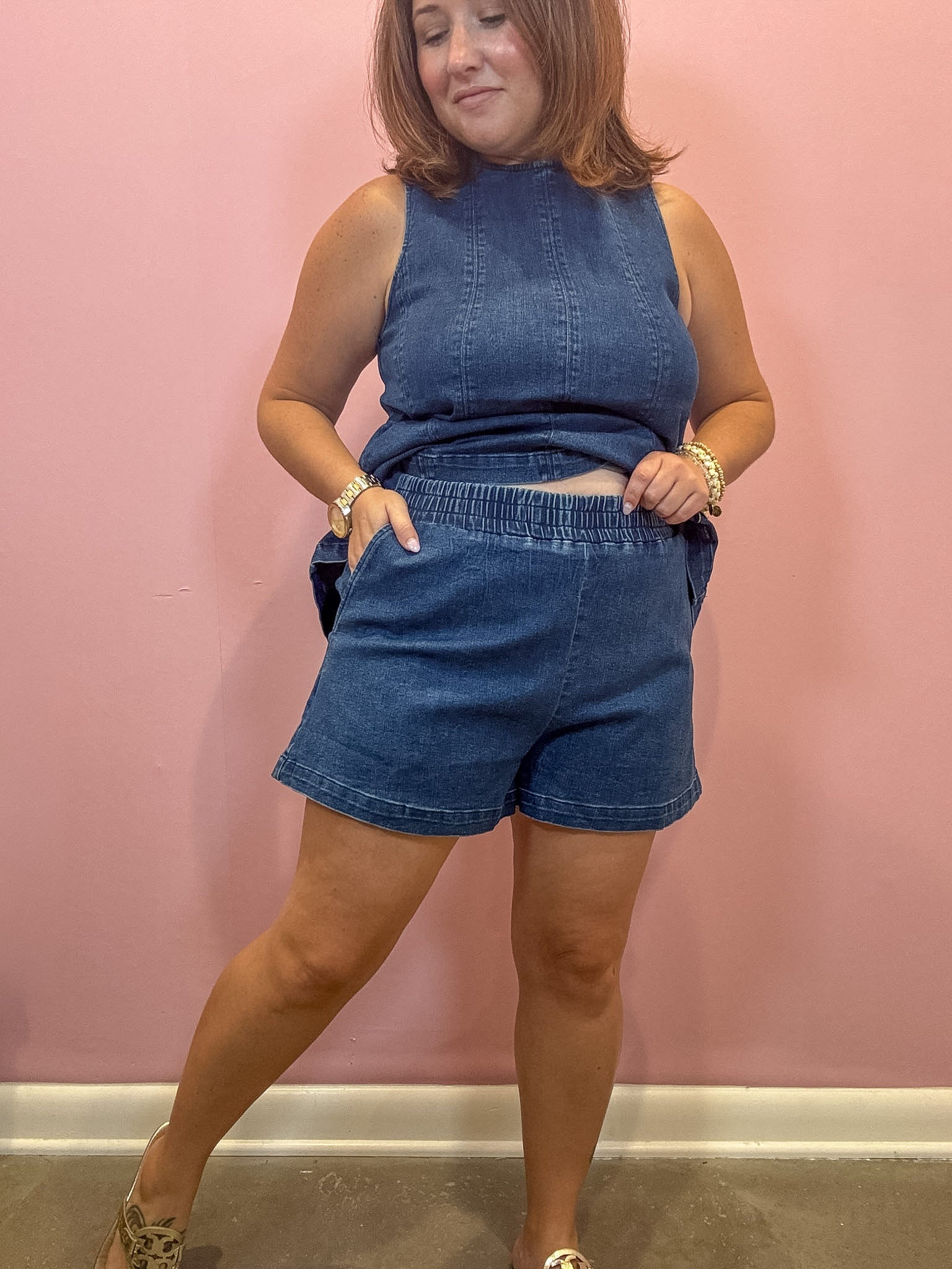 A woman modeling a matching two-piece denim set consisting of a sleeveless high-neck top and elastic-waist shorts, posed against a solid pink wall.