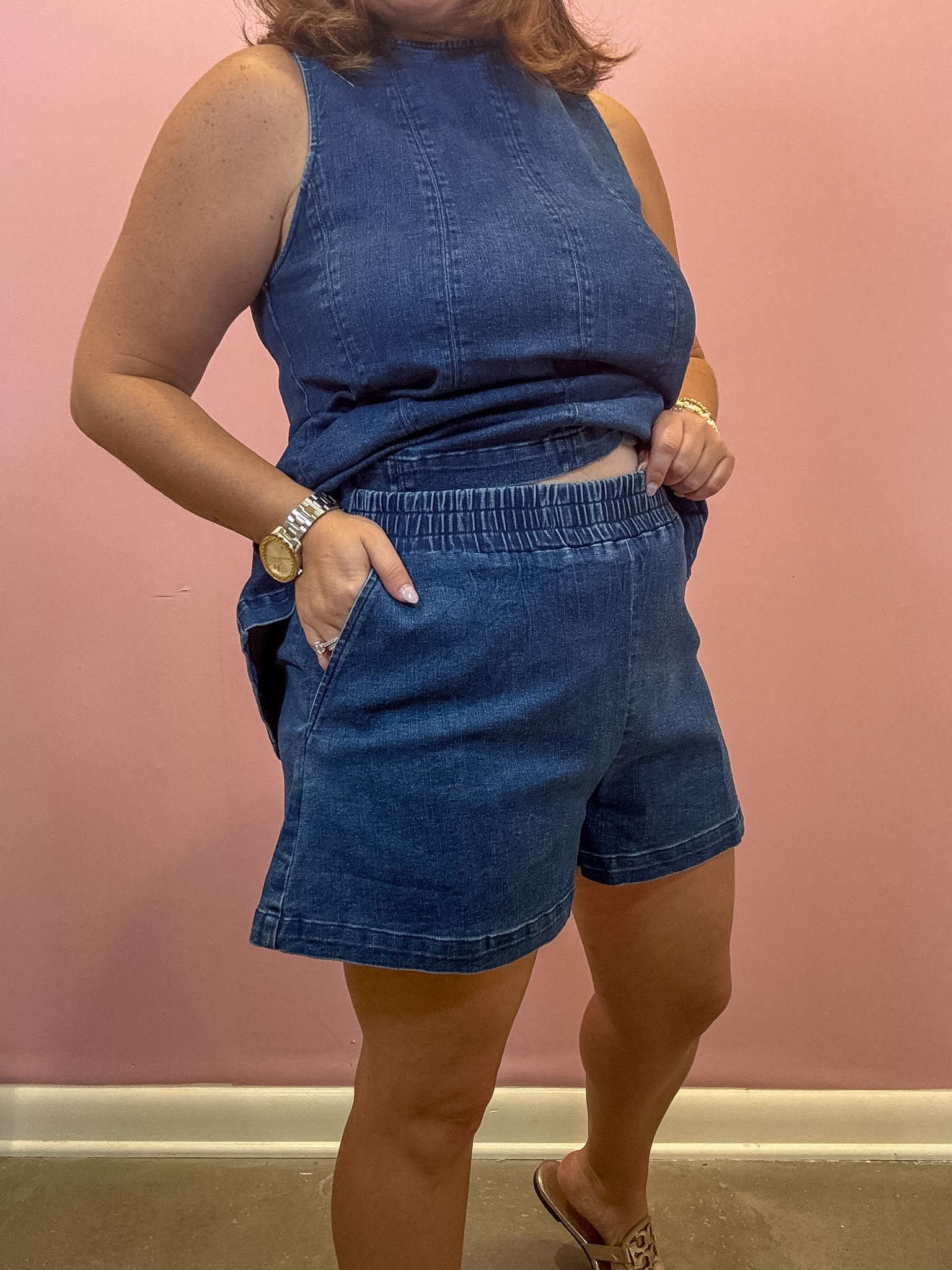 A woman modeling a matching blue denim set consisting of a sleeveless top and high-waisted shorts, standing against a pink wall.