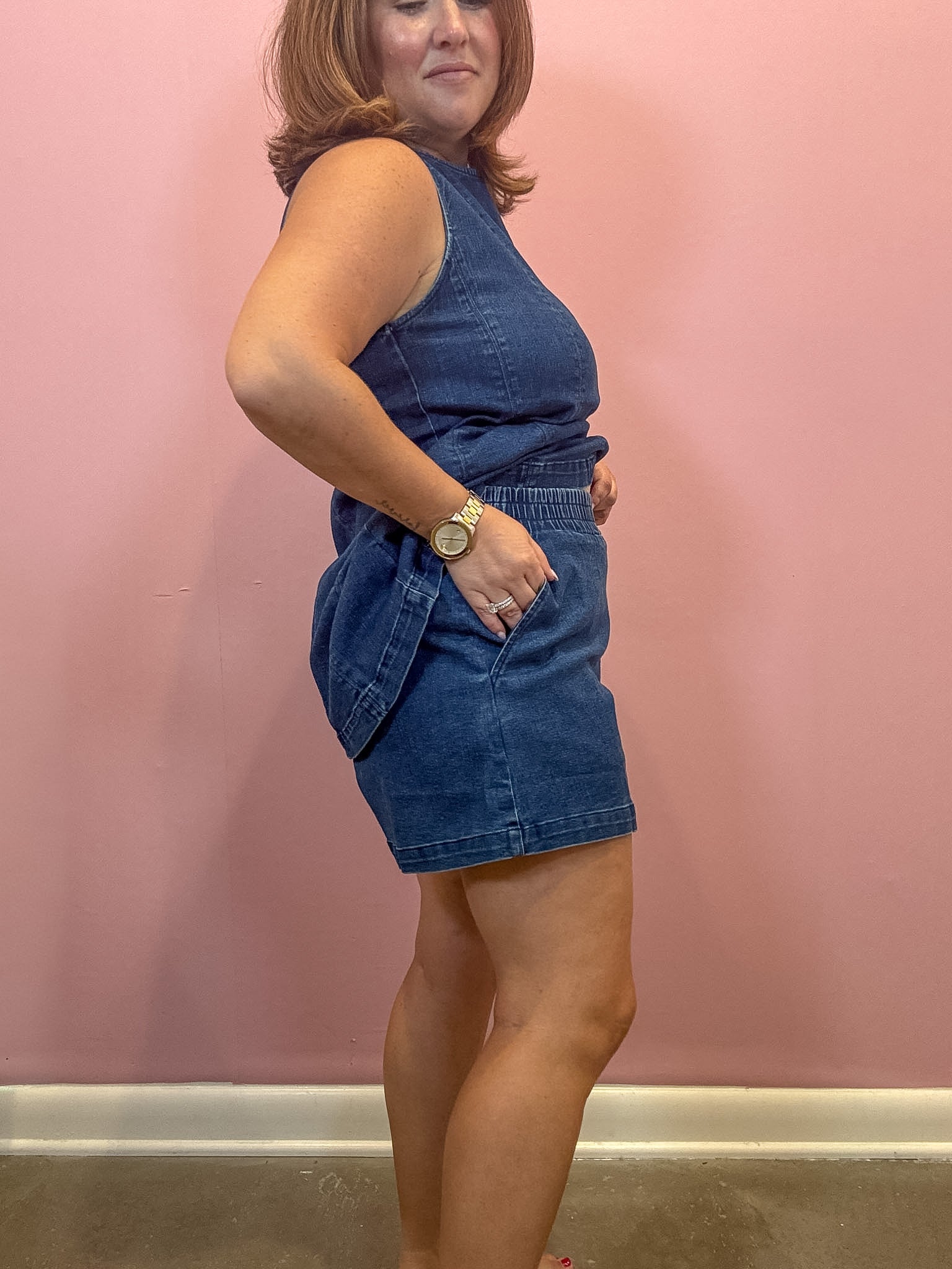 A woman modeling a sleeveless blue denim romper against a pink wall.