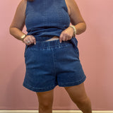 A woman modeling a matching blue denim two-piece set consisting of a sleeveless top and high-waisted shorts, standing against a pink wall.