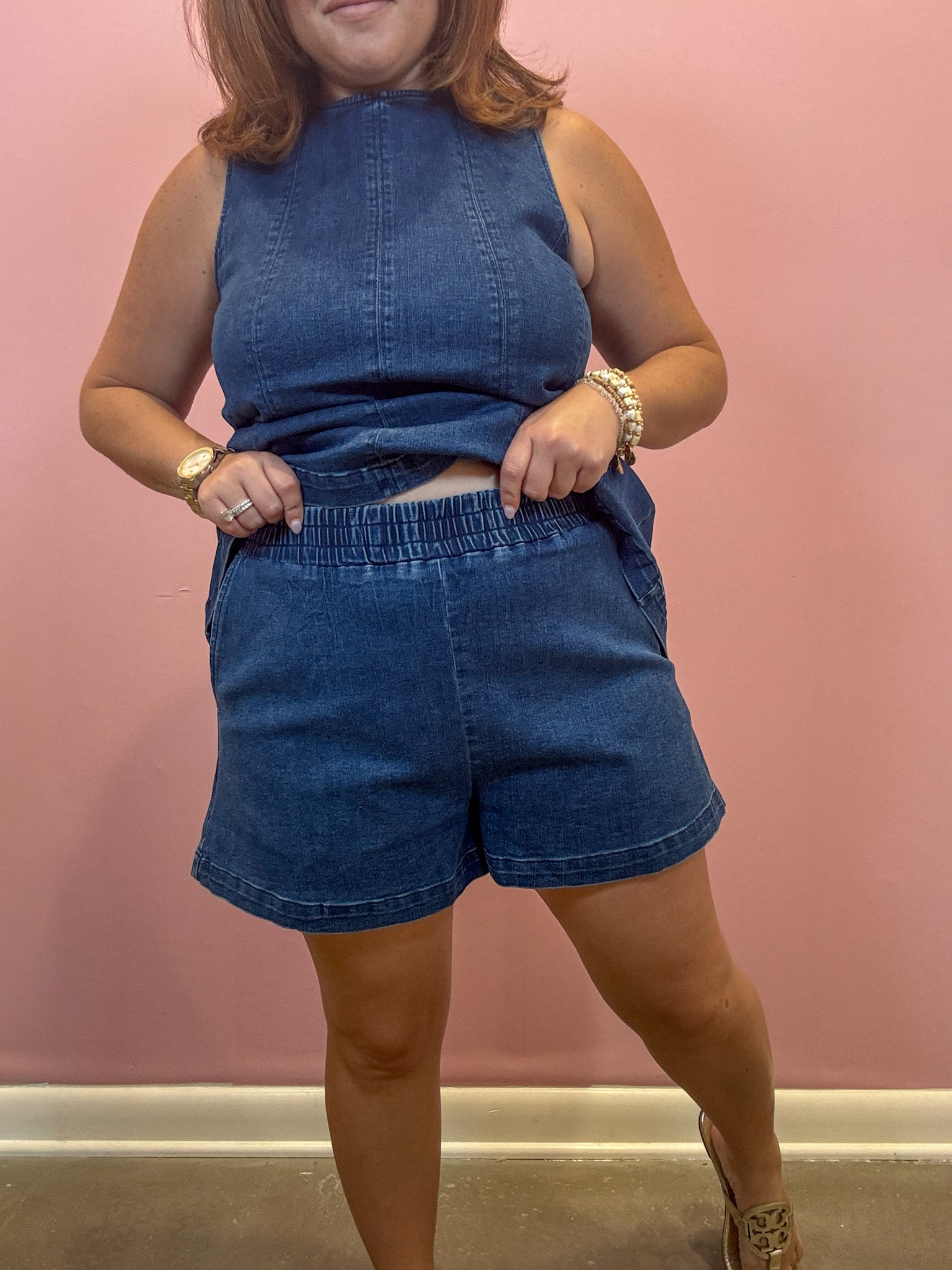 A woman modeling a matching blue denim two-piece set consisting of a sleeveless top and high-waisted shorts, standing against a pink wall.
