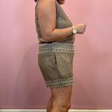 A woman modeling a sleeveless olive green linen romper with white embroidered trim, standing against a pink wall.