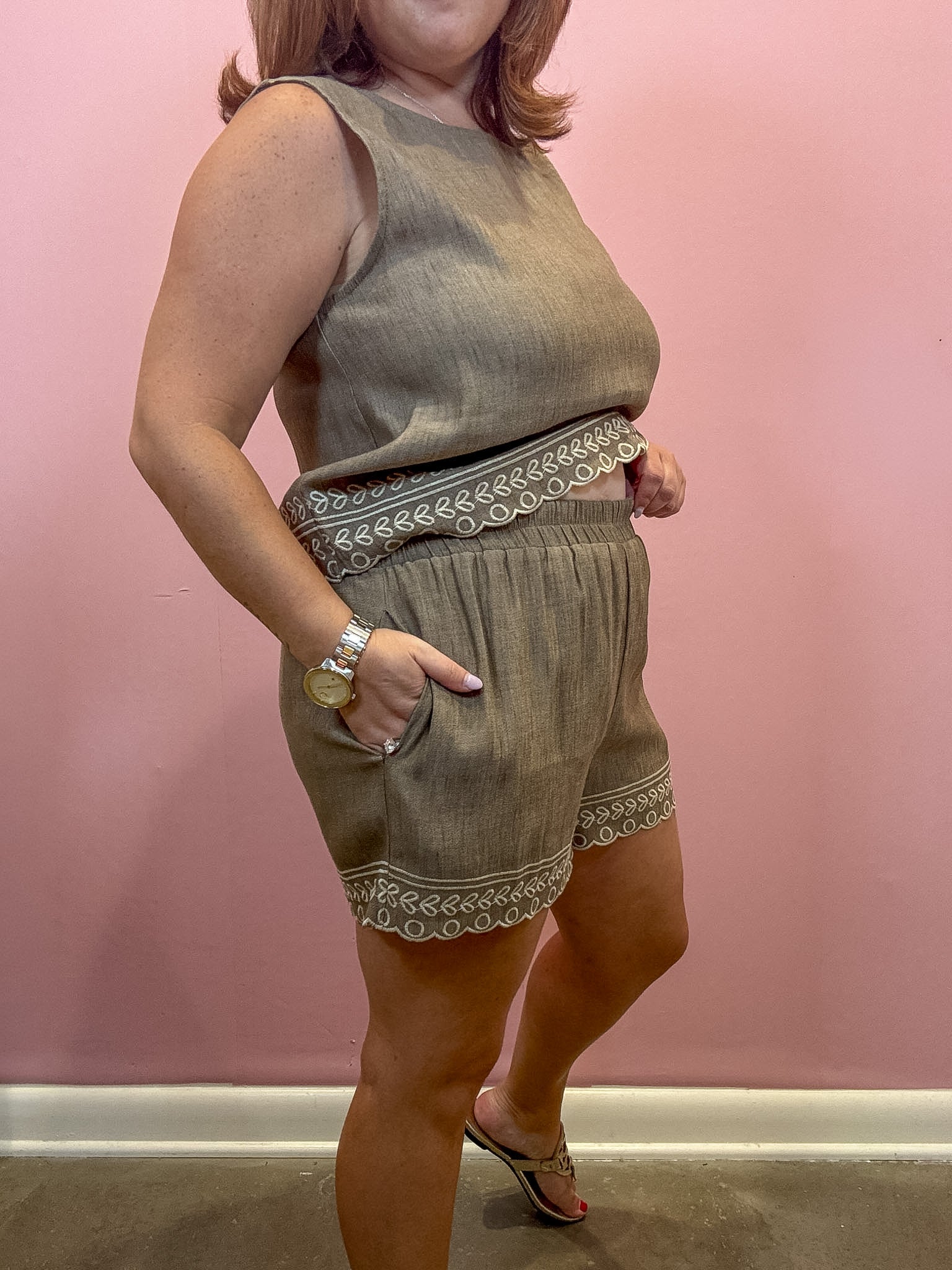 A woman modeling a sleeveless olive green linen romper with white embroidered trim, standing against a pink wall.