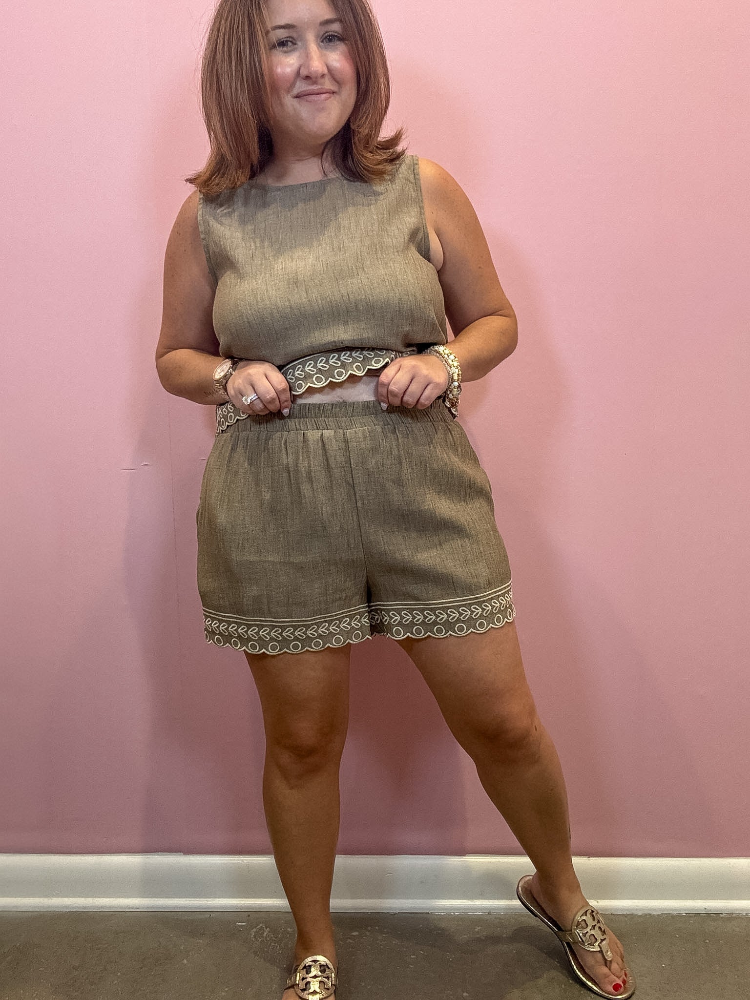 A woman modeling a sleeveless taupe linen romper with decorative lace trim, standing against a pink wall.