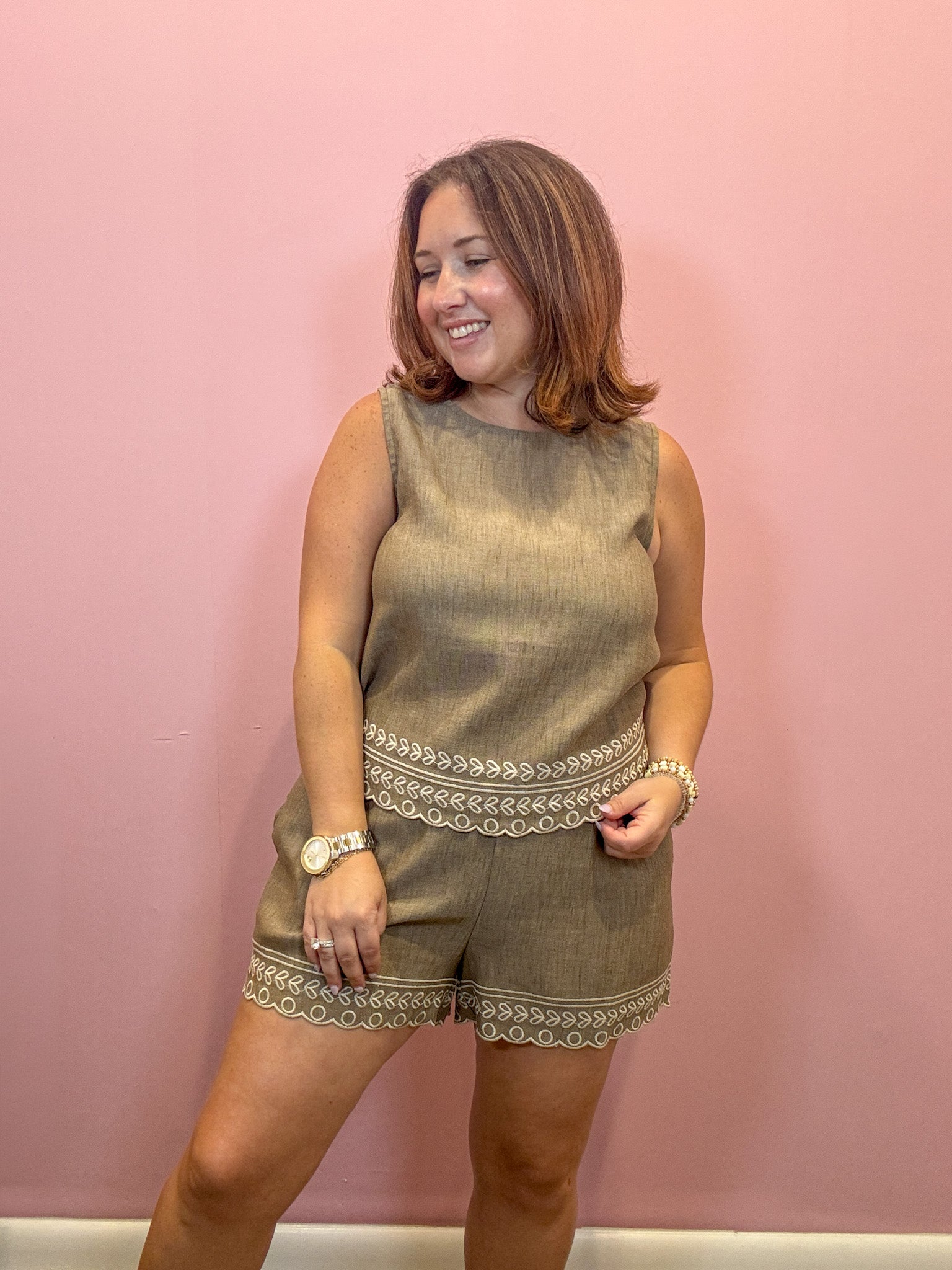 A woman modeling a sleeveless olive green linen romper with beige embroidered trim, standing against a solid pink wall.