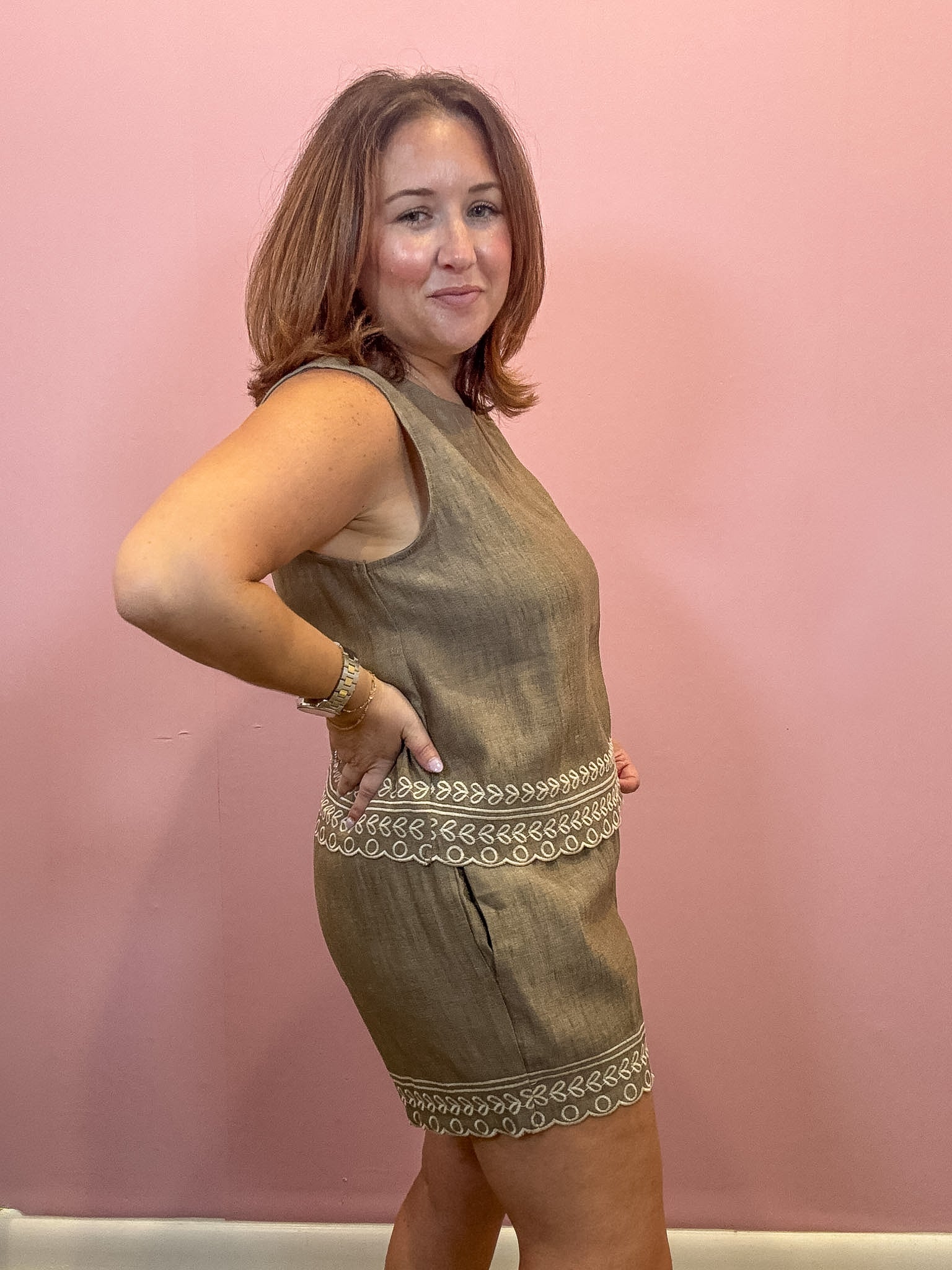 A woman modeling a sleeveless olive green linen mini dress with decorative embroidered trim at the waist and hem, standing against a solid pink wall.