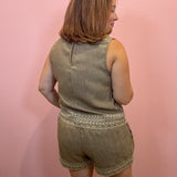 A woman shown from the back wearing a sleeveless olive-brown linen romper with embroidered trim, standing against a solid pink wall.