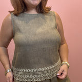 A woman wearing a sleeveless olive green linen jumpsuit with a decorative embroidered border at the waist, standing against a solid pink wall.