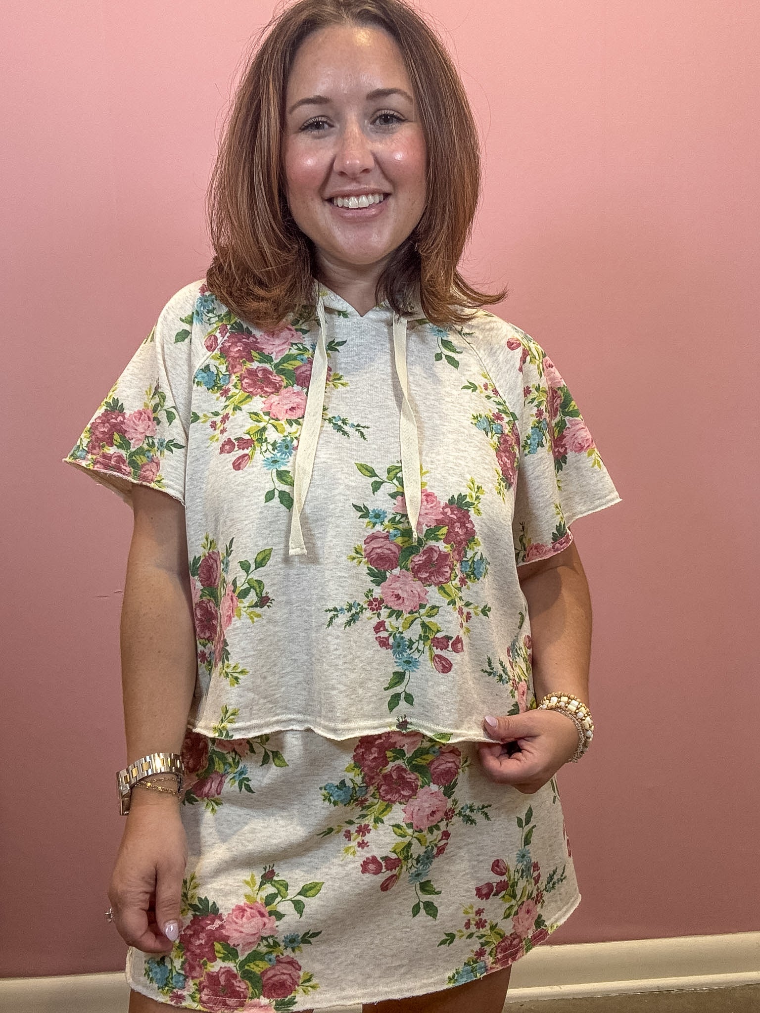 A woman wearing a short-sleeve floral print hoodie and matching mini skirt set, standing against a solid pink wall.