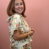 A woman with shoulder-length brown hair smiling and looking over her shoulder, wearing a matching two-piece floral loungewear set consisting of a short-sleeve top and shorts. The set has a light beige base with a pink, green, and blue floral pattern, set against a solid dusty pink background.