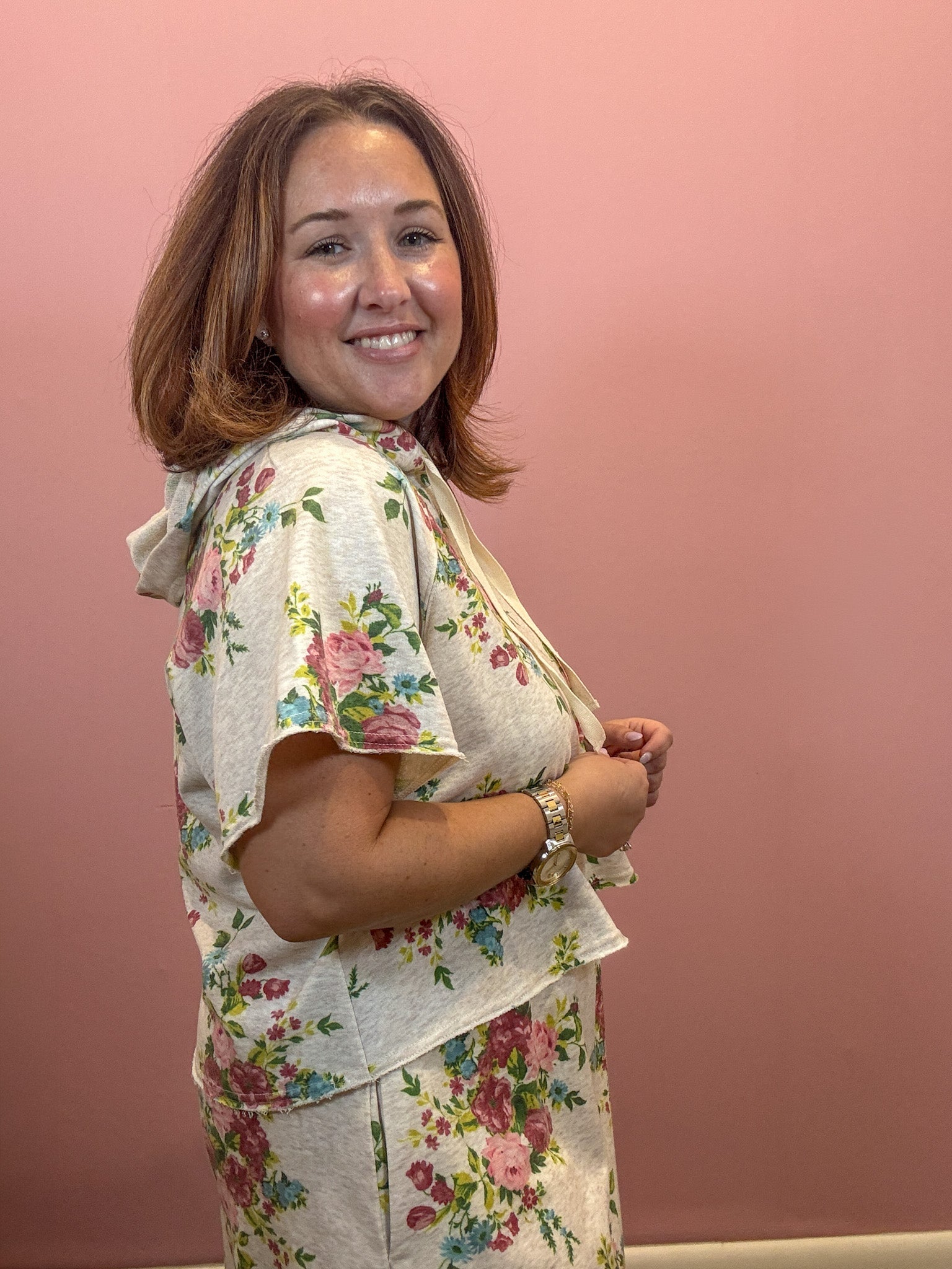 A woman with shoulder-length brown hair smiling and looking over her shoulder, wearing a matching two-piece floral loungewear set consisting of a short-sleeve top and shorts. The set has a light beige base with a pink, green, and blue floral pattern, set against a solid dusty pink background.
