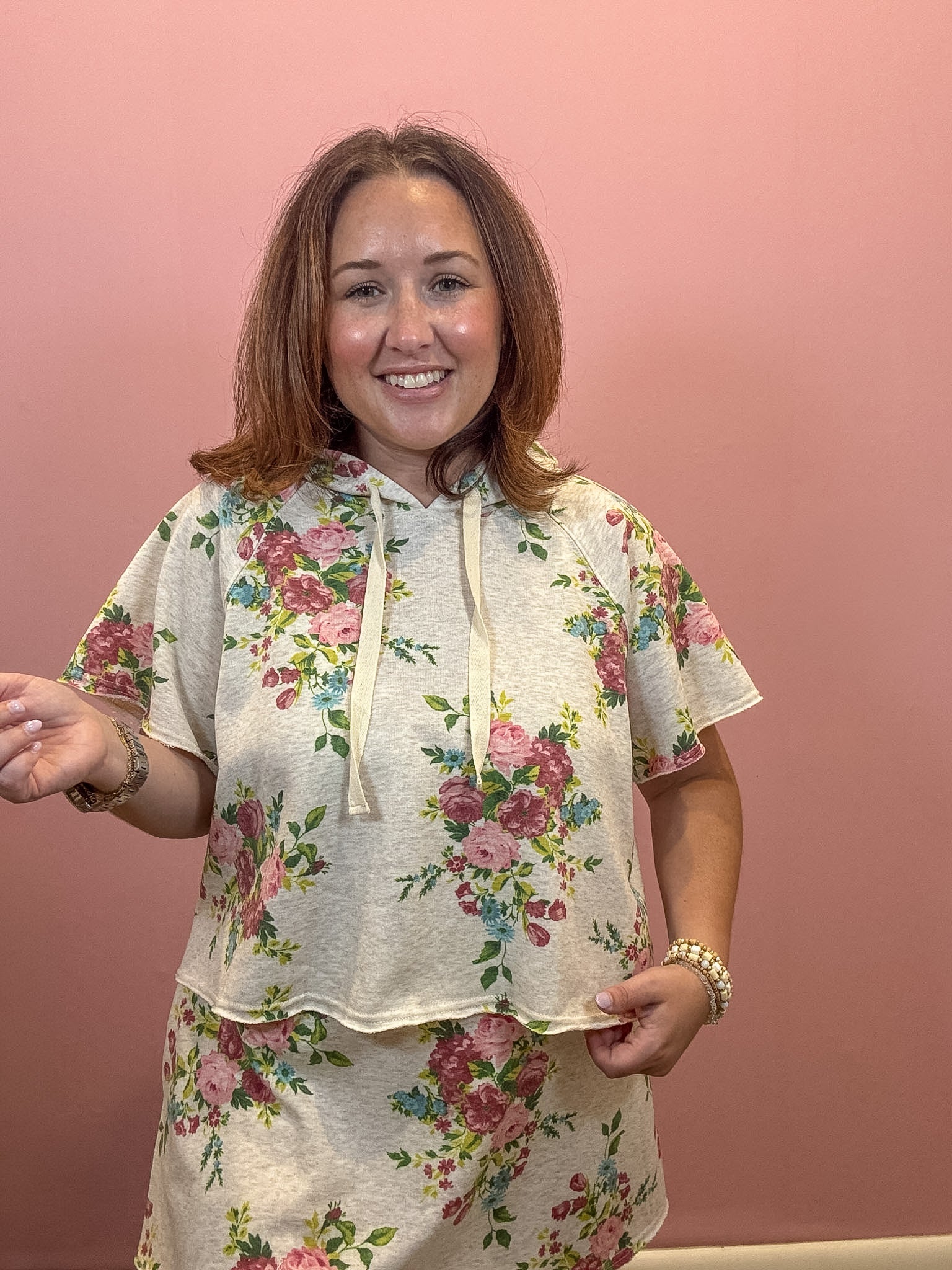 A woman wearing a matching two-piece floral lounge set consisting of a short-sleeve hooded top and a matching skirt, set against a solid pink wall.
