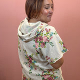 A woman wearing a light beige hooded short-sleeve top with a pink, green, and blue floral print, shown from a side-back angle against a solid pink wall.