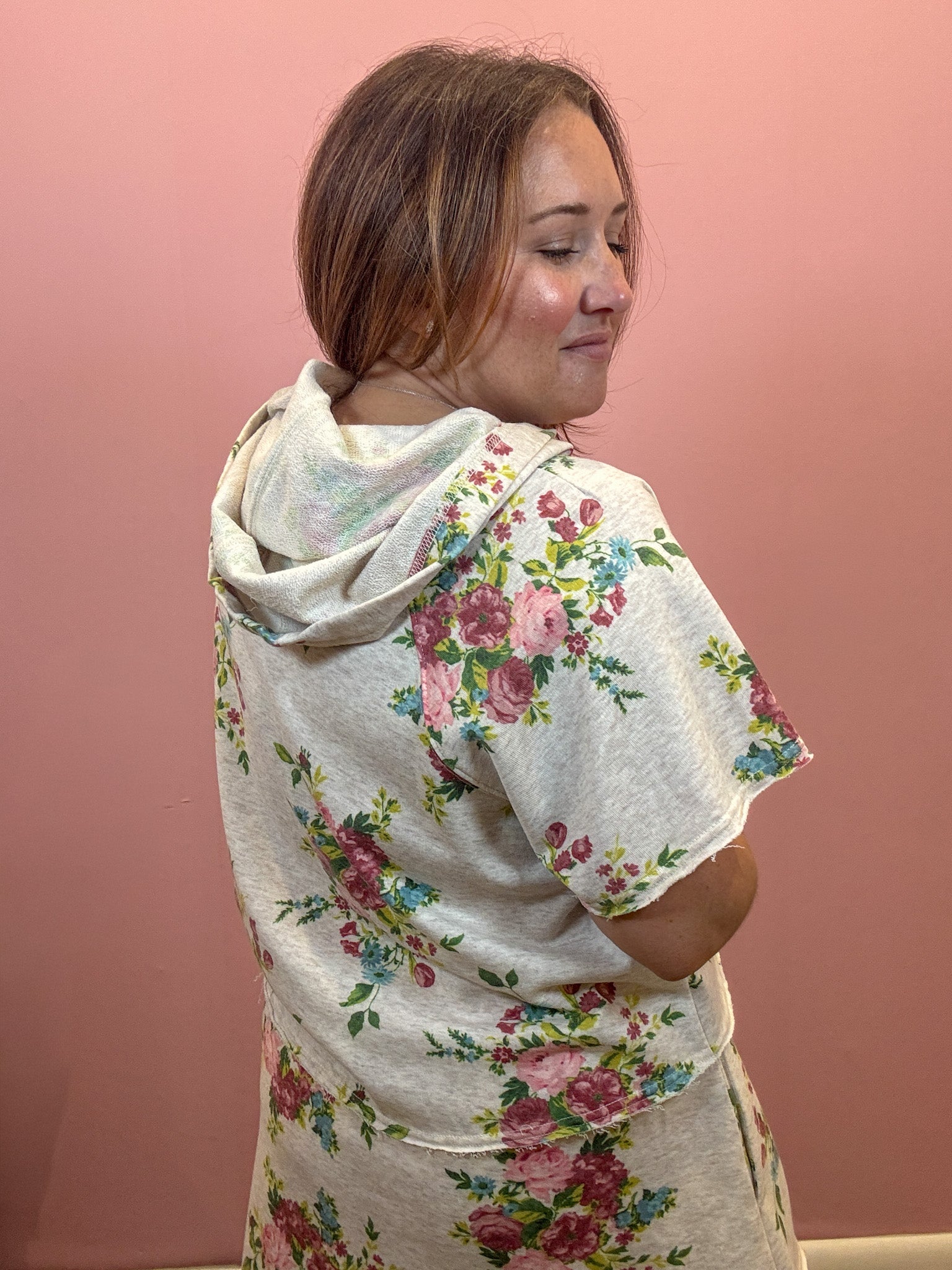 A woman wearing a light beige hooded short-sleeve top with a pink, green, and blue floral print, shown from a side-back angle against a solid pink wall.