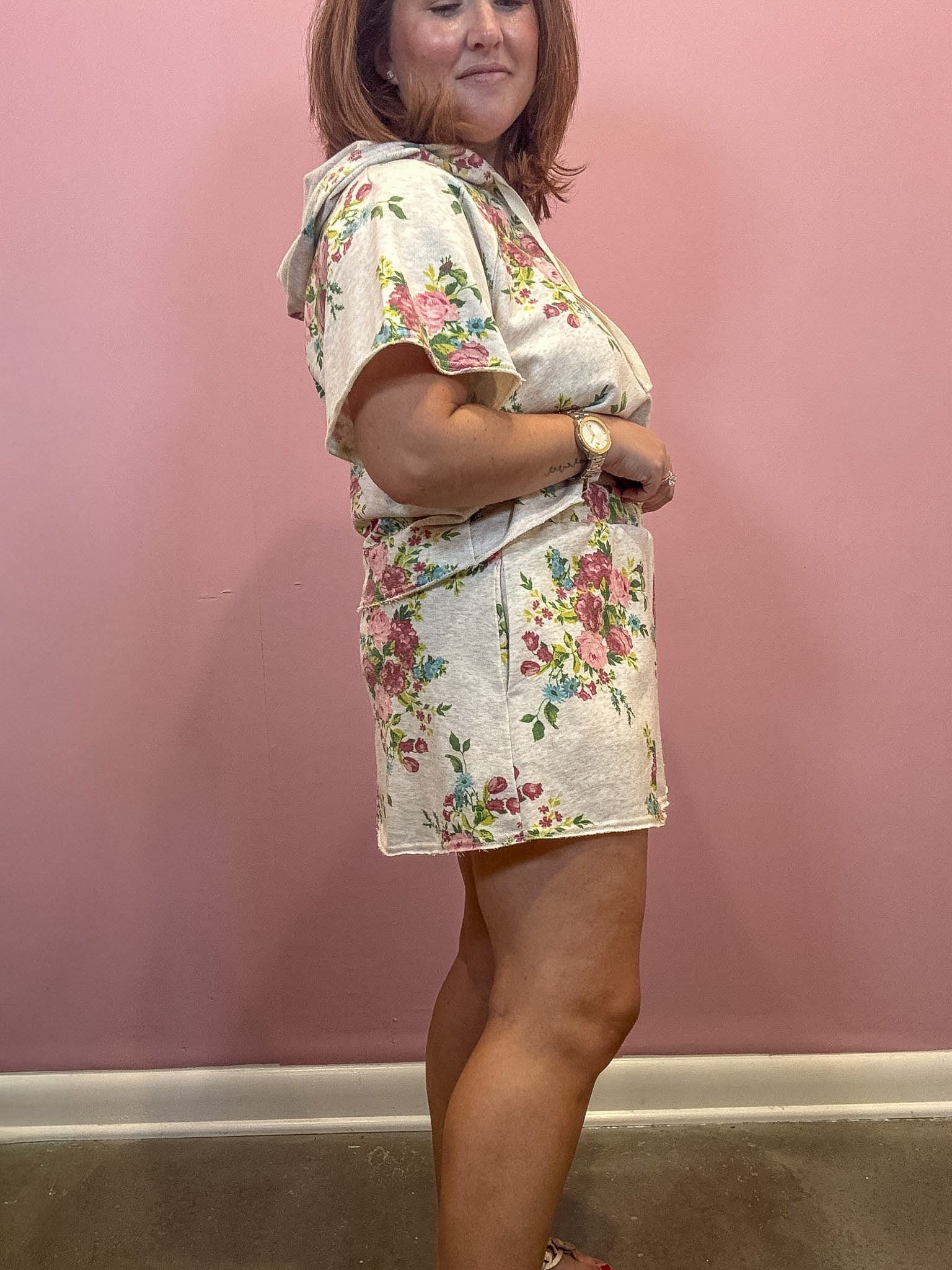 A woman modeling a matching short-sleeve top and mini skirt set with a pink and green floral pattern on a light beige background, standing against a dusty pink wall.