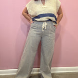 A woman modeling a cream knit sleeveless top with a navy blue horizontal stripe and wide-leg gray pinstriped pants with a cream drawstring, standing against a pink wall.