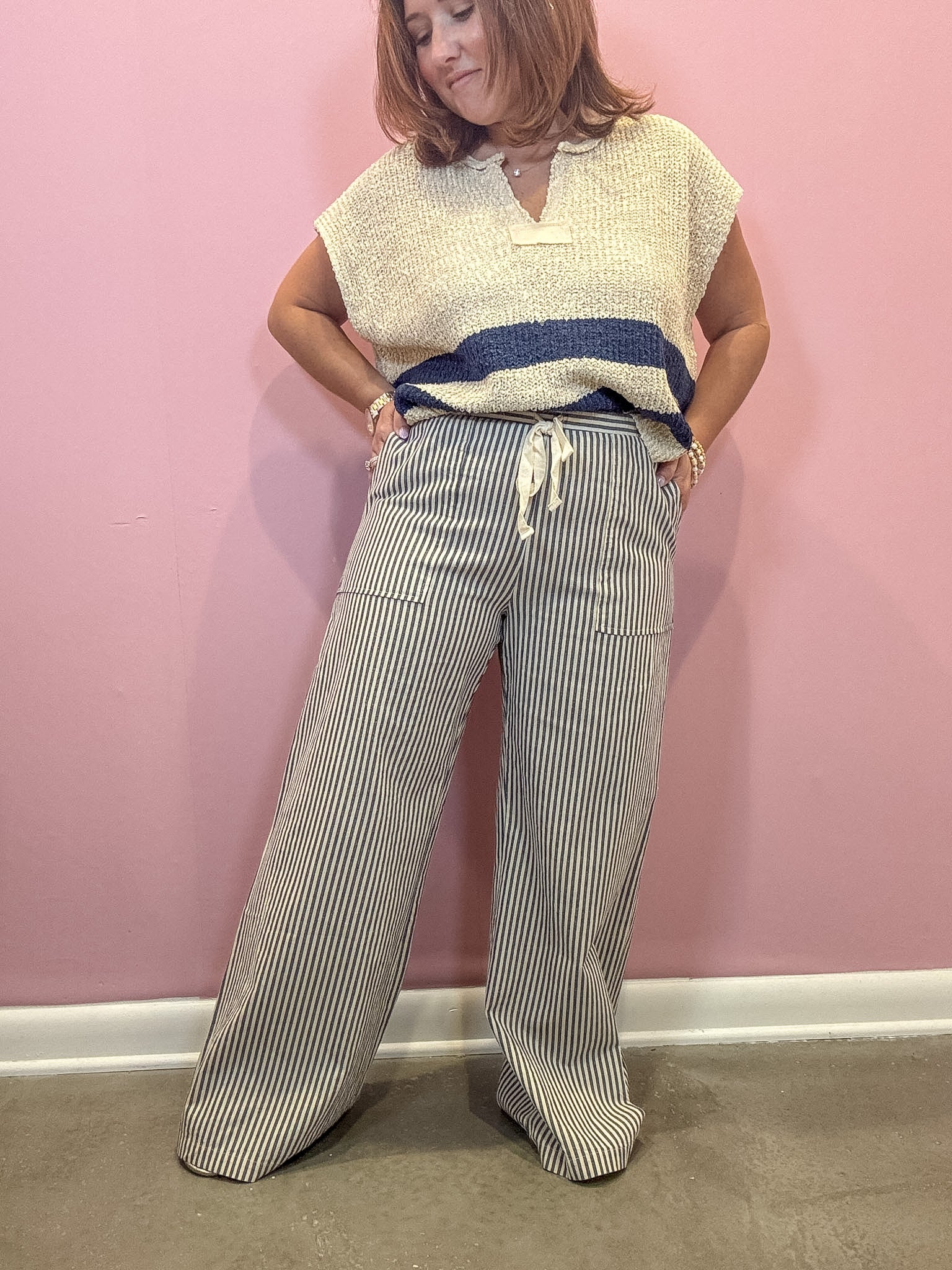 A woman modeling a cream knit sleeveless top with a navy blue horizontal stripe and wide-leg gray pinstriped pants with a cream drawstring, standing against a pink wall.