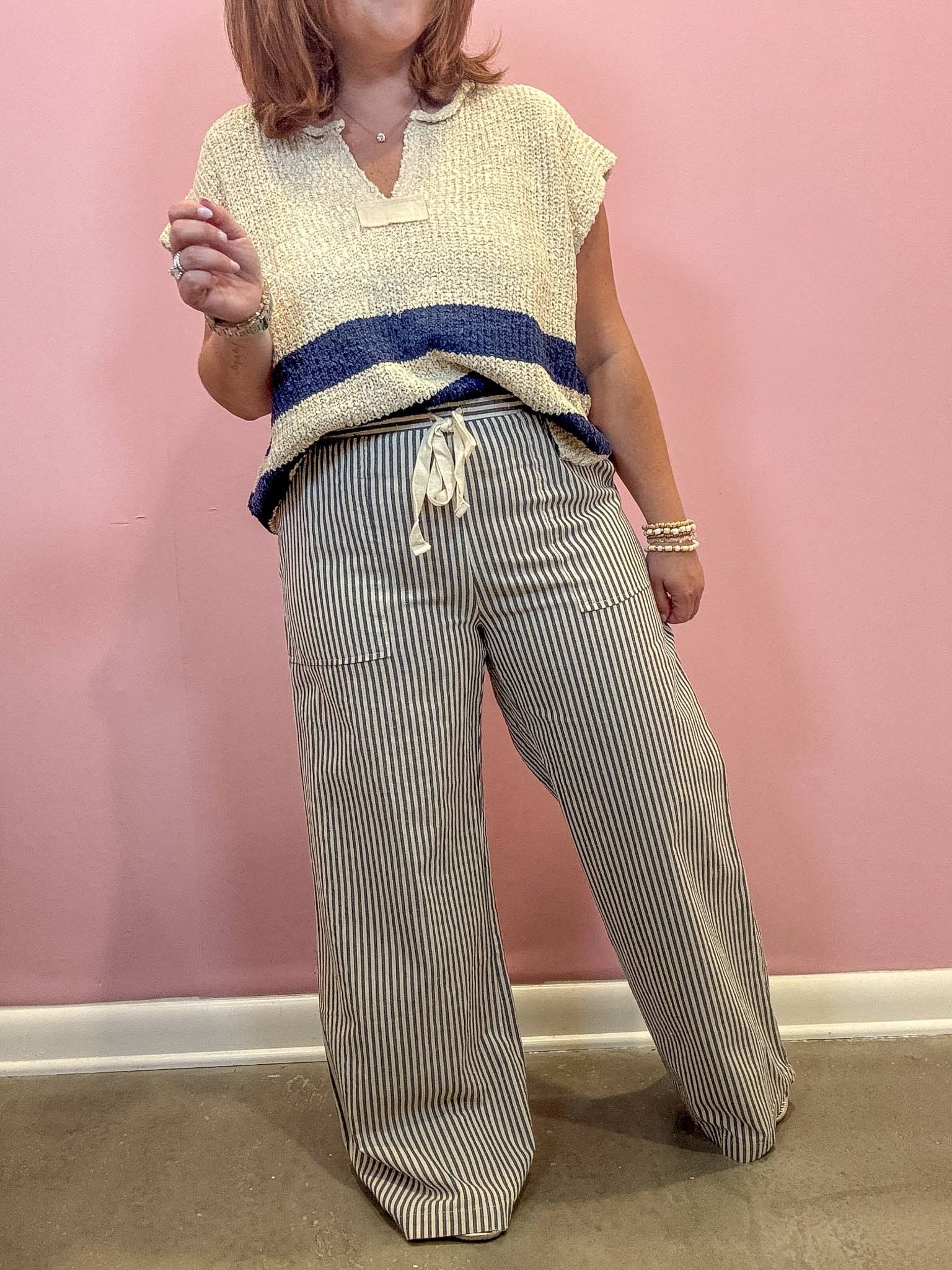 A person wearing a cream knit sleeveless top with a navy blue horizontal stripe and striped wide-leg pants, standing against a pink wall.