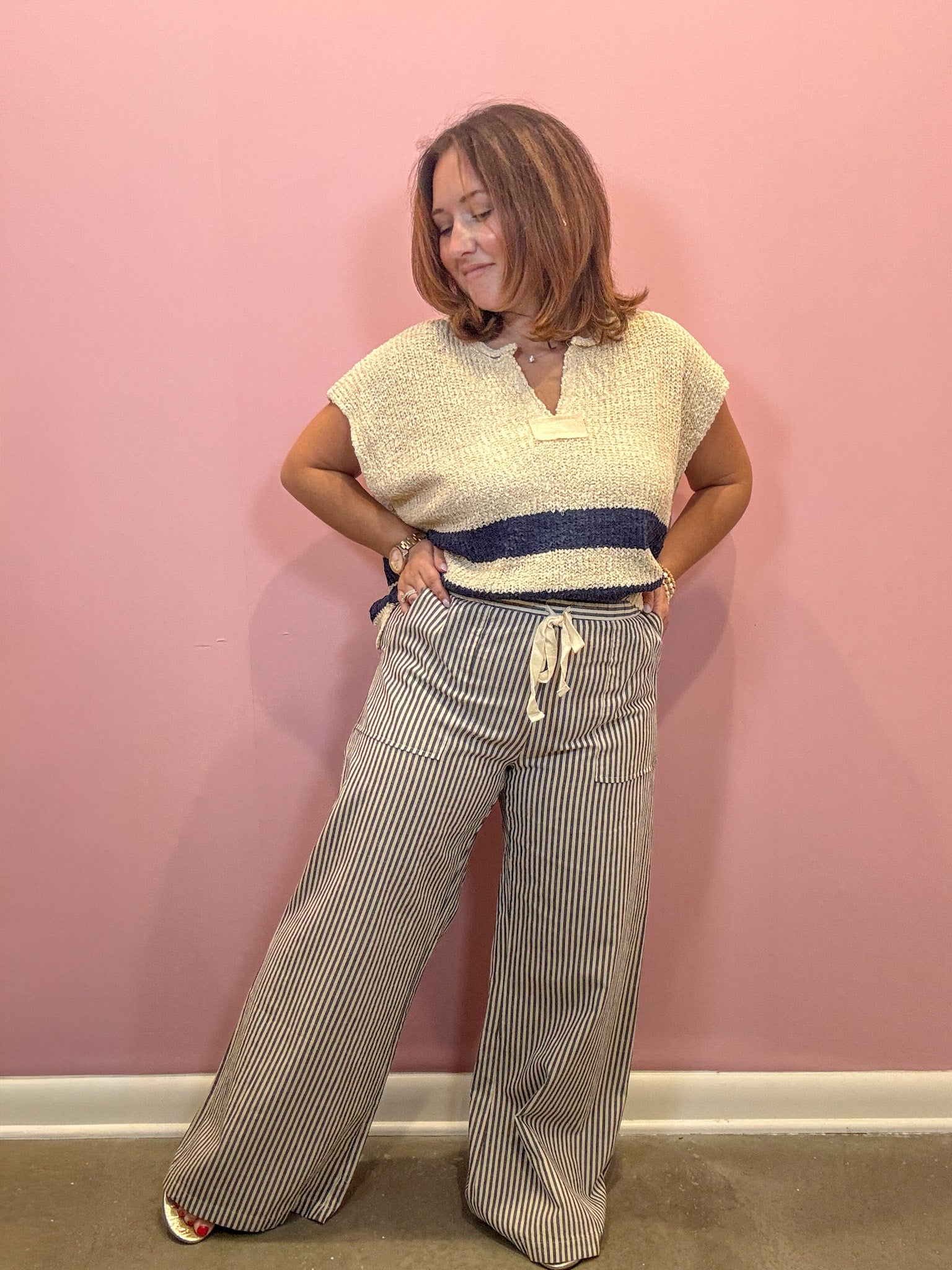 A woman posing in a cream knit short-sleeve top with a navy blue horizontal stripe and matching striped wide-leg pants, standing against a solid pink wall.