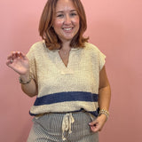 A woman smiling and posing in a textured cream knit top with a navy blue horizontal stripe and striped pants, set against a solid pink wall.
