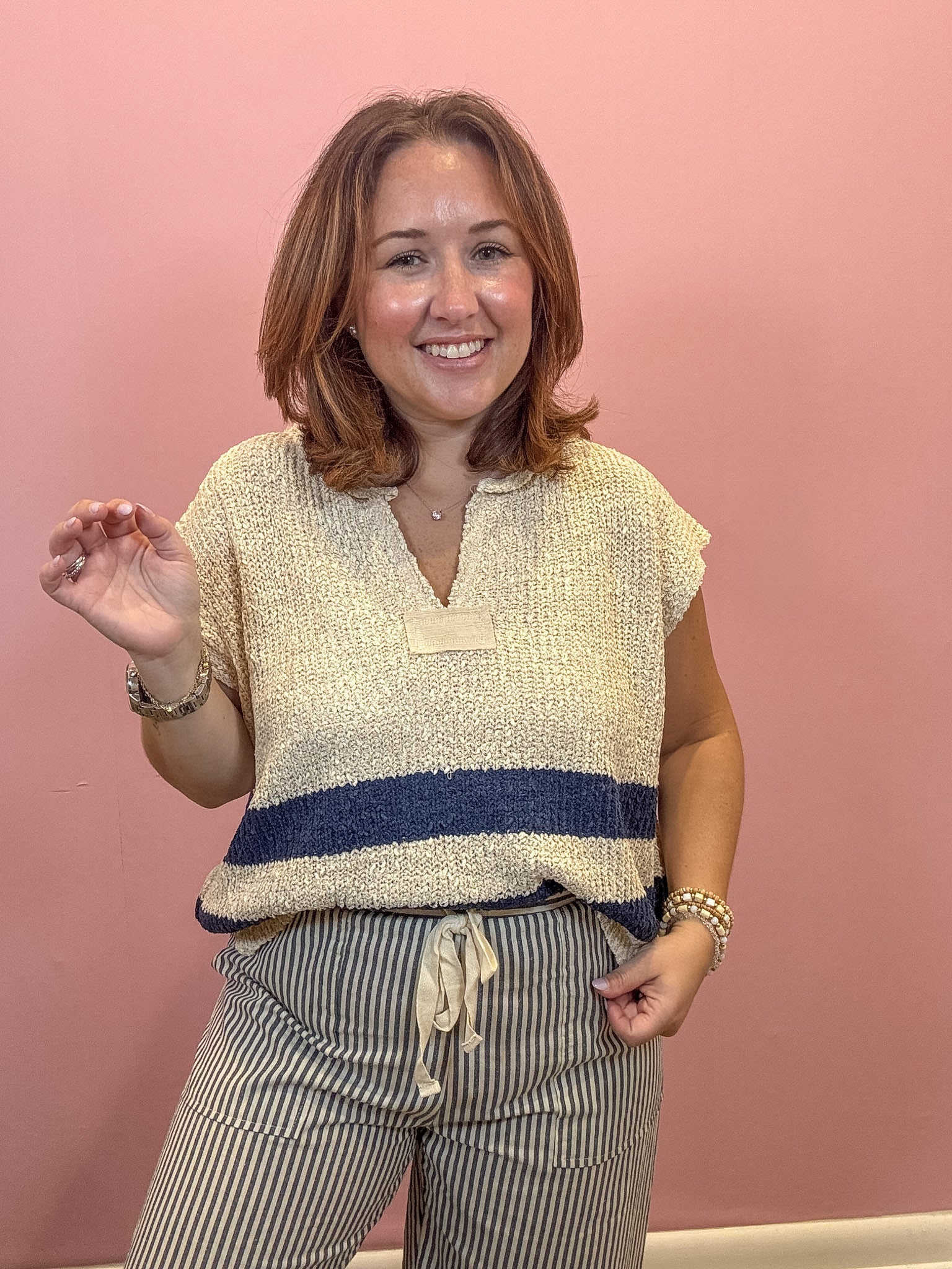 A woman smiling and posing in a textured cream knit top with a navy blue horizontal stripe and striped pants, set against a solid pink wall.
