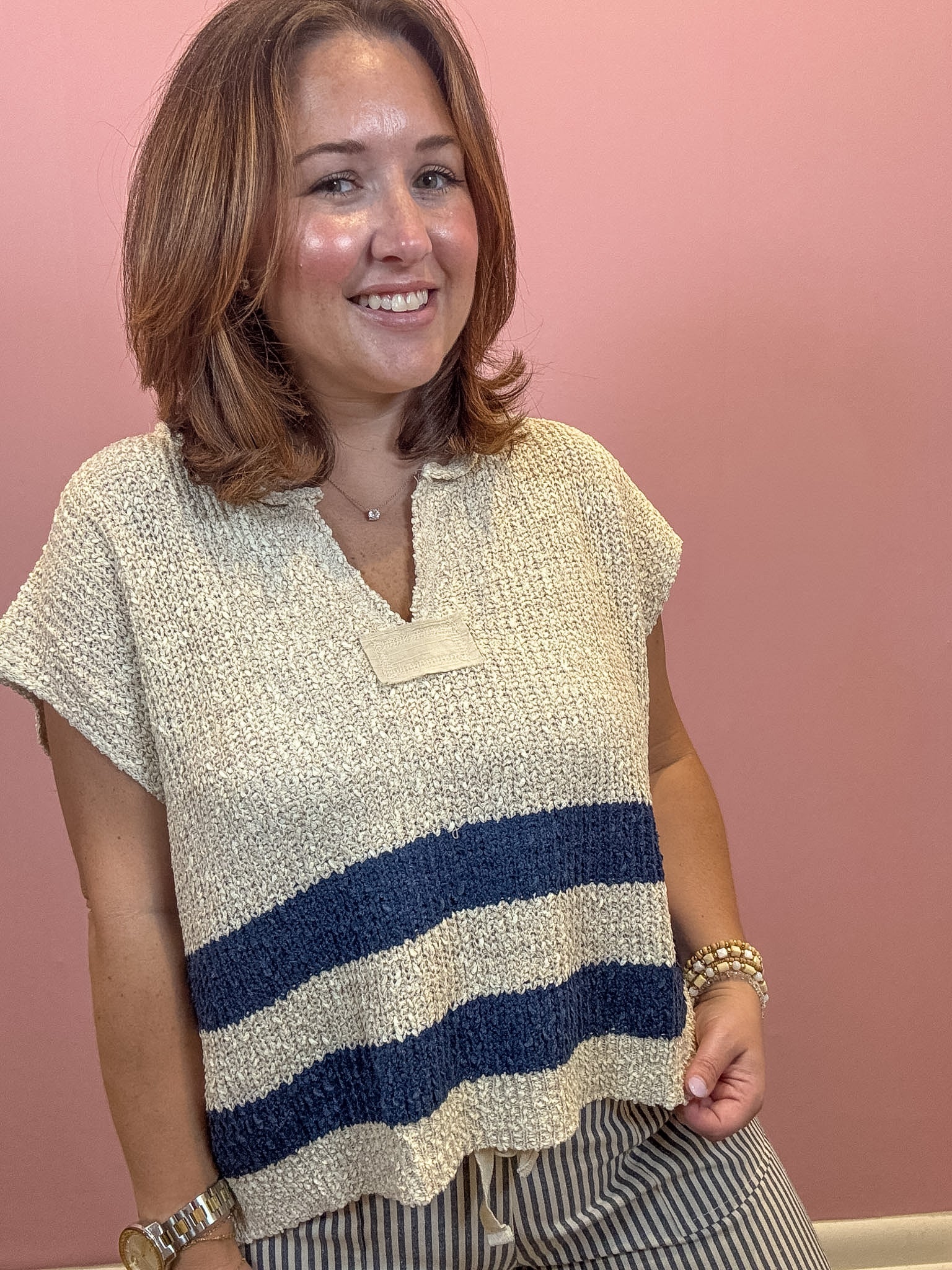 A woman wearing a textured knit top with a cream base and navy blue horizontal stripes, standing against a solid pink wall.