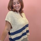 A woman wearing a cream and navy blue striped knit poncho top, paired with striped pants, standing against a solid pink wall.