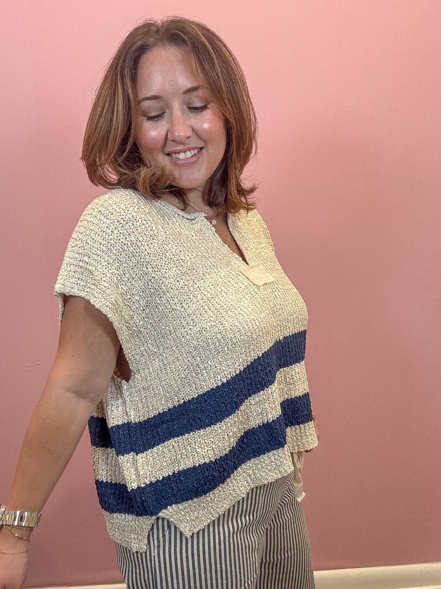 A woman wearing a cream and navy blue striped knit poncho top, paired with striped pants, standing against a solid pink wall.