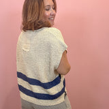 A woman with shoulder-length brown hair is shown from the back, wearing a cream-colored textured knit sweater with two horizontal navy blue stripes near the bottom, paired with gray and white vertically striped pants, set against a solid pink wall.