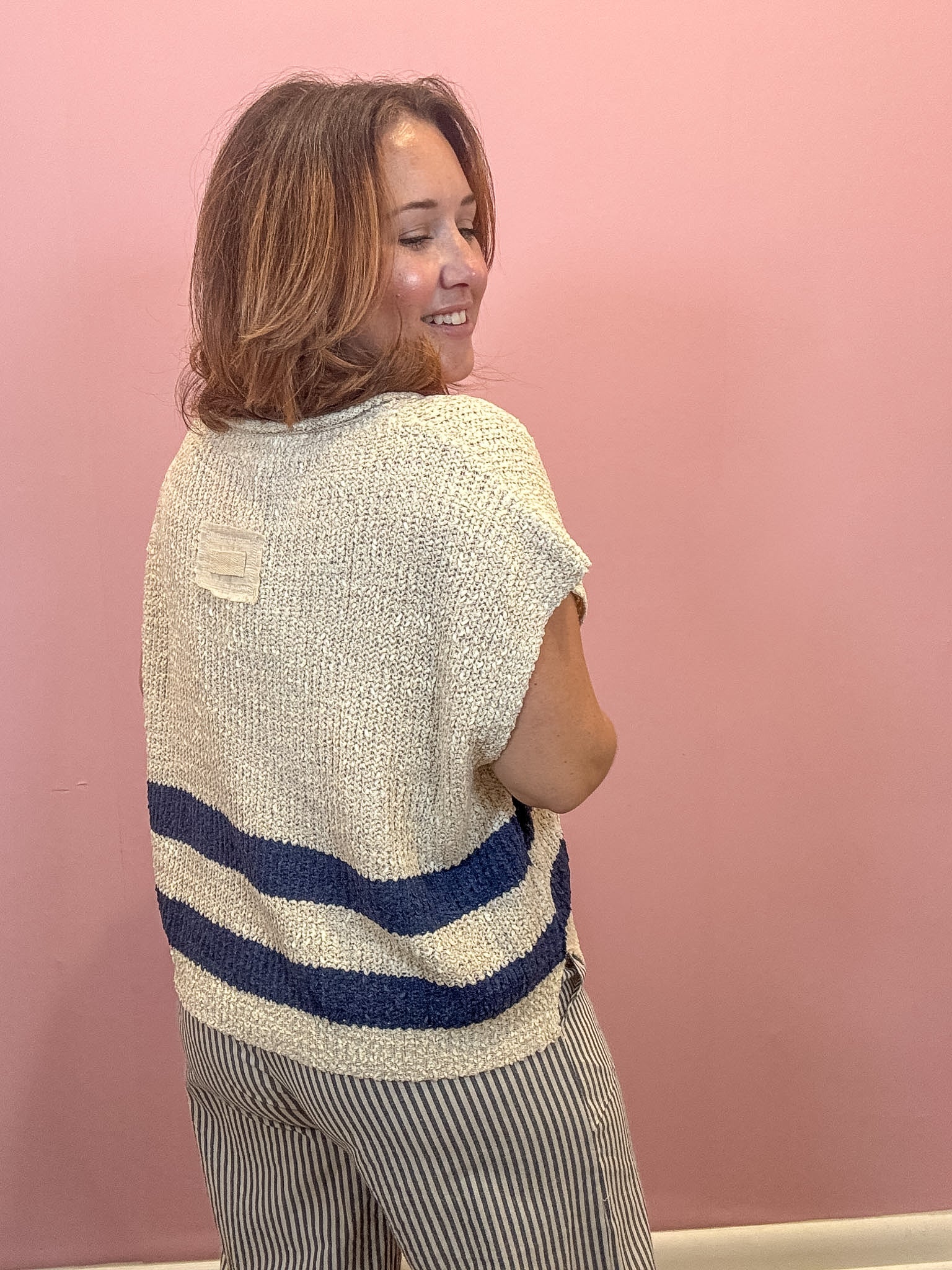 A woman with shoulder-length brown hair is shown from the back, wearing a cream-colored textured knit sweater with two horizontal navy blue stripes near the bottom, paired with gray and white vertically striped pants, set against a solid pink wall.