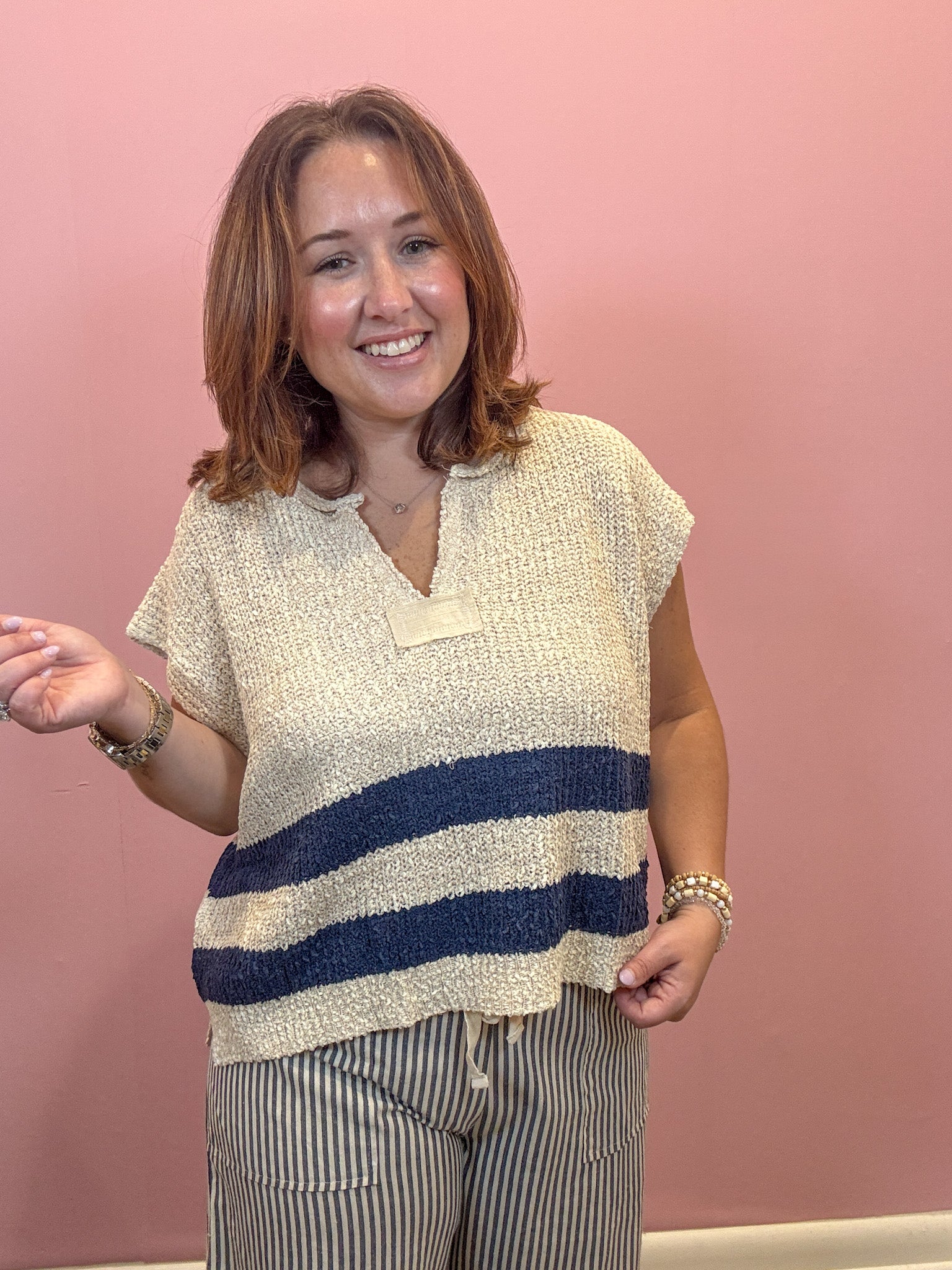 A woman wearing a textured knit top with a cream base and navy blue horizontal stripes, paired with striped pants, standing against a solid pink wall.
