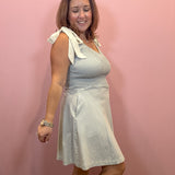 A woman modeling a sleeveless A-line dress with diagonal stripes in light blue and white, featuring a white bow detail on the shoulder, standing against a solid pink wall.