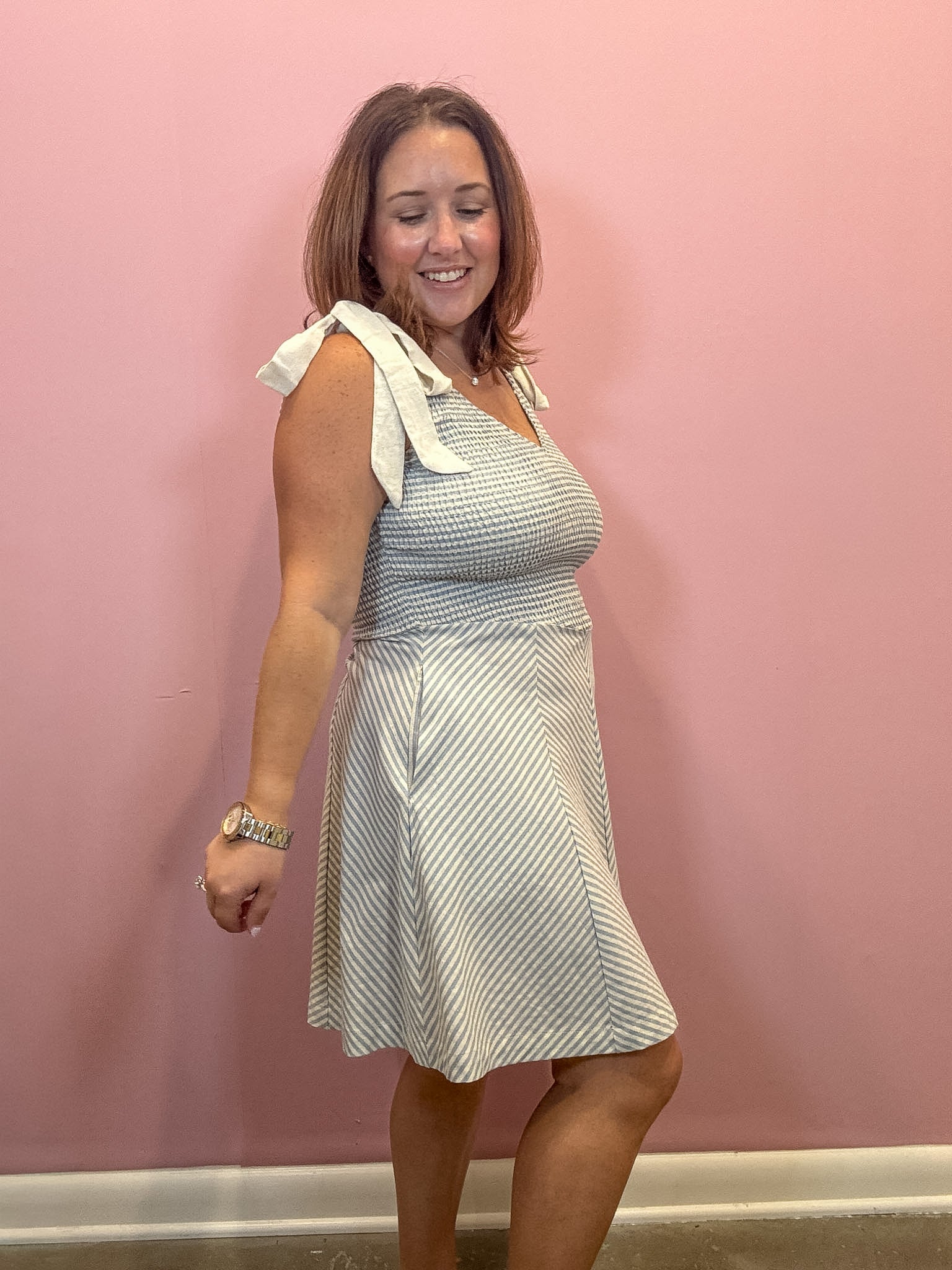 A woman modeling a sleeveless A-line dress with diagonal stripes in light blue and white, featuring a white bow detail on the shoulder, standing against a solid pink wall.