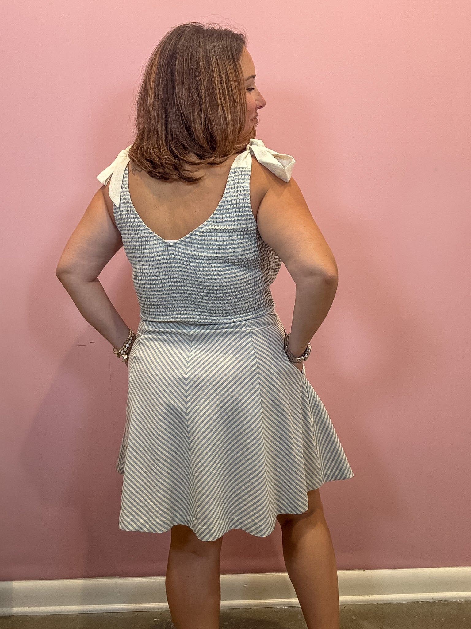 A woman shown from the back wearing a sleeveless smocked dress with white bow details on the shoulders, set against a solid pink wall.