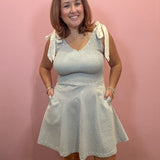 A woman modeling a sleeveless V-neck dress with tie shoulder straps and side pockets, featuring a textured blue and white pattern, standing against a solid pink wall.
