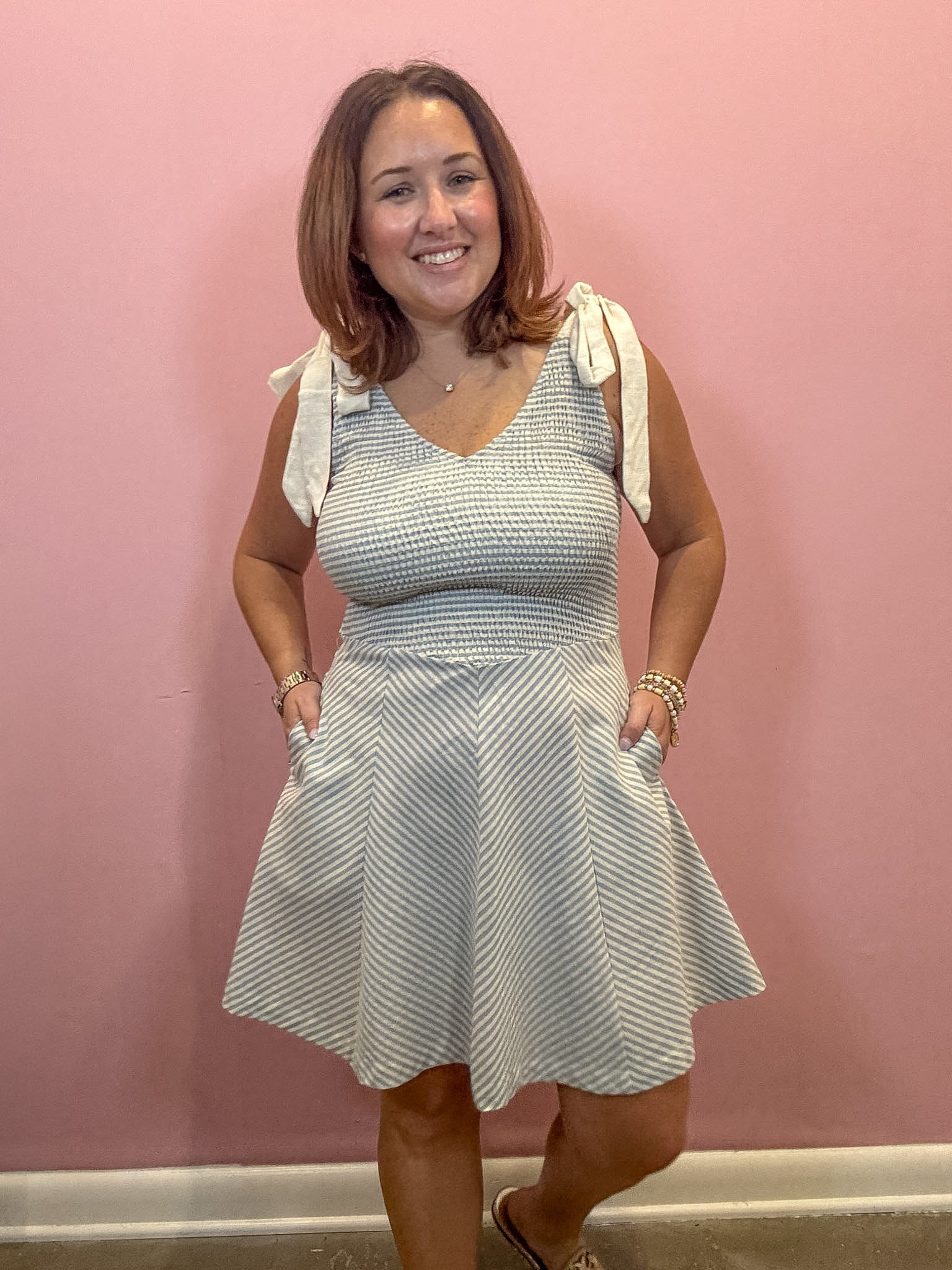 A woman modeling a sleeveless V-neck dress with tie shoulder straps and side pockets, featuring a textured blue and white pattern, standing against a solid pink wall.