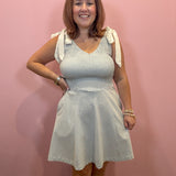 A woman modeling a sleeveless V-neck dress with a textured chevron pattern in light blue and white, featuring white tie details on the shoulders, standing against a pink wall.