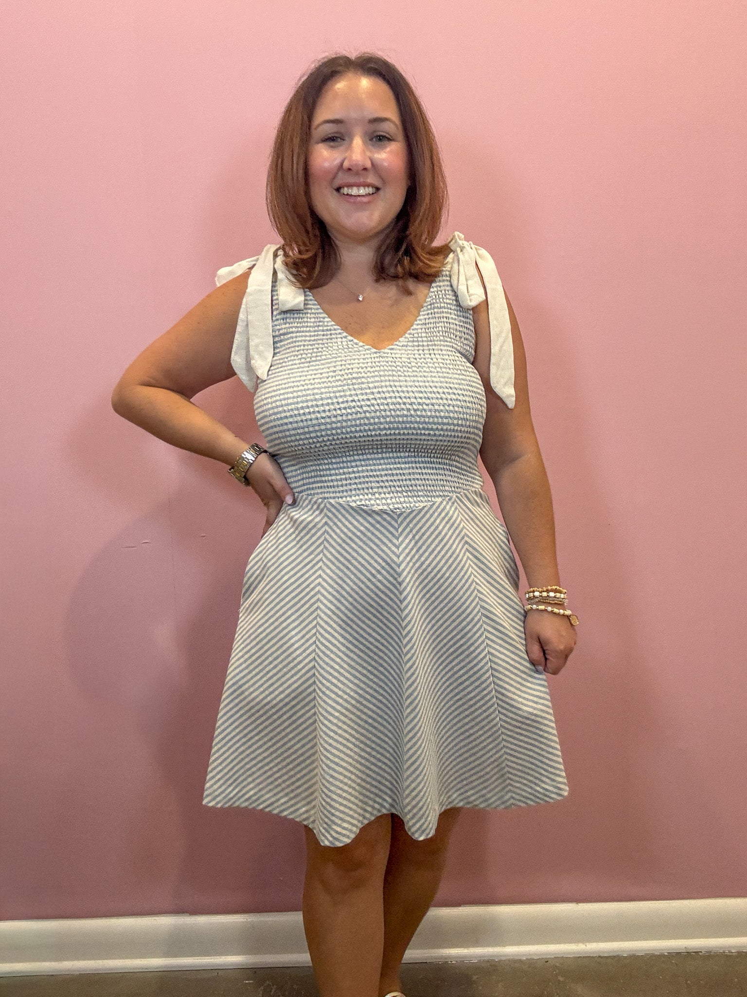A woman modeling a sleeveless V-neck dress with a textured chevron pattern in light blue and white, featuring white tie details on the shoulders, standing against a pink wall.