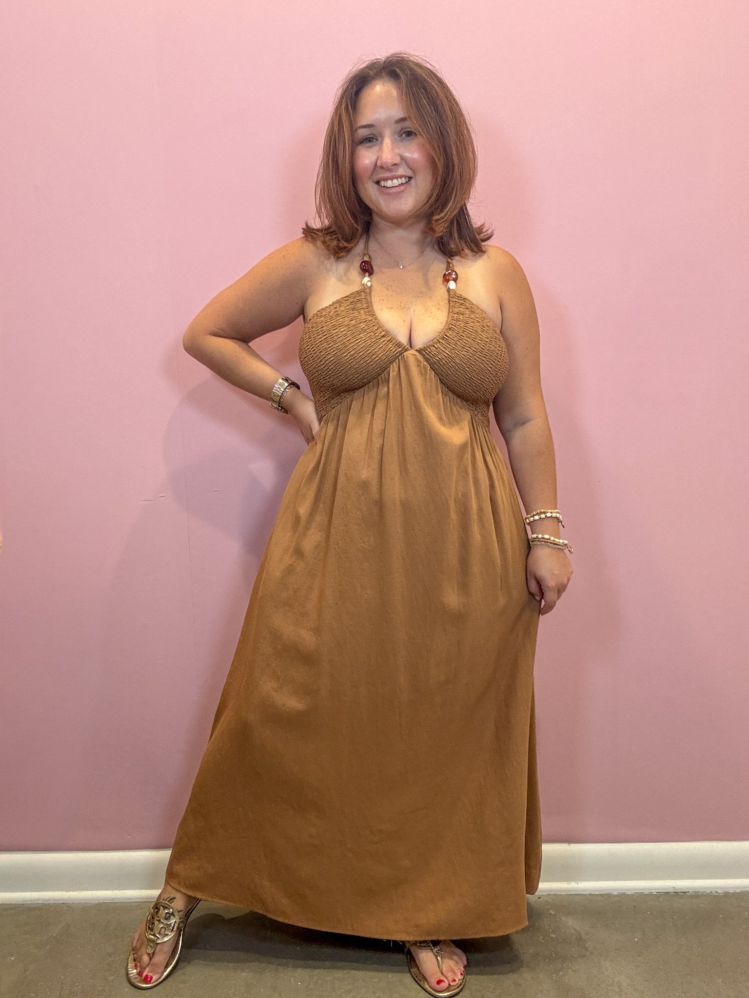 A woman modeling a brown halter-neck maxi dress with a textured bodice and flowing skirt, standing against a solid pink wall.