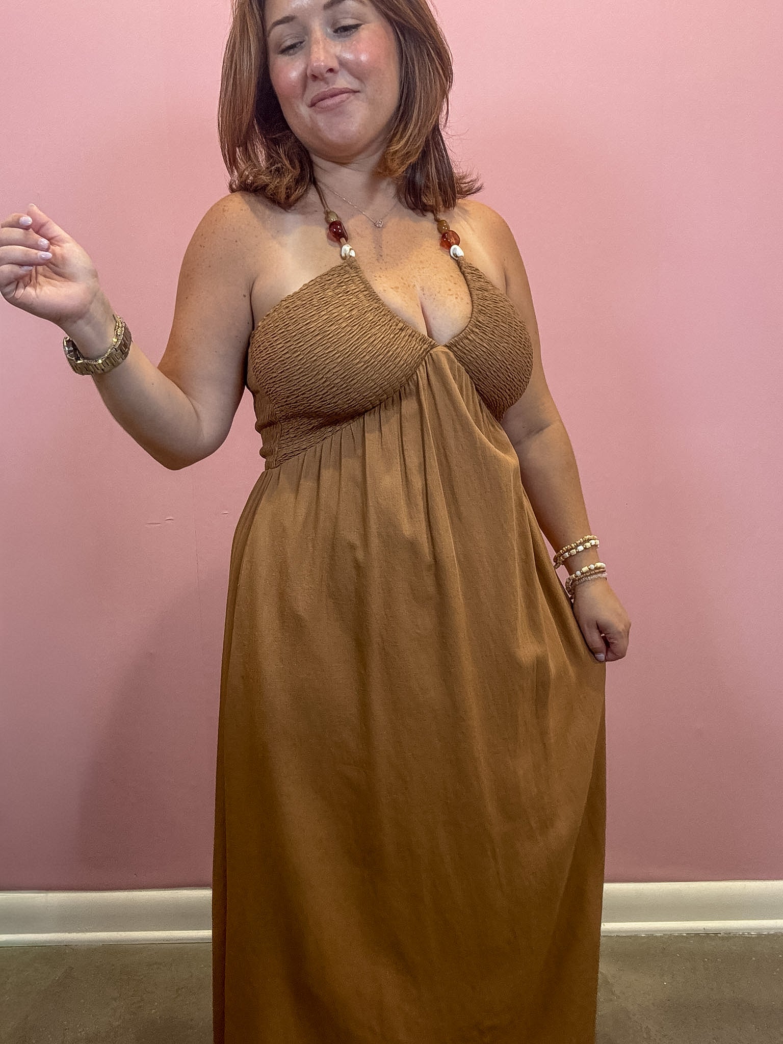 A woman modeling a brown halter-neck maxi dress with a smocked bodice, standing against a solid pink wall.