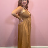 A woman modeling a sleeveless, smocked-bust maxi dress in a warm camel brown color, standing against a solid pink wall.