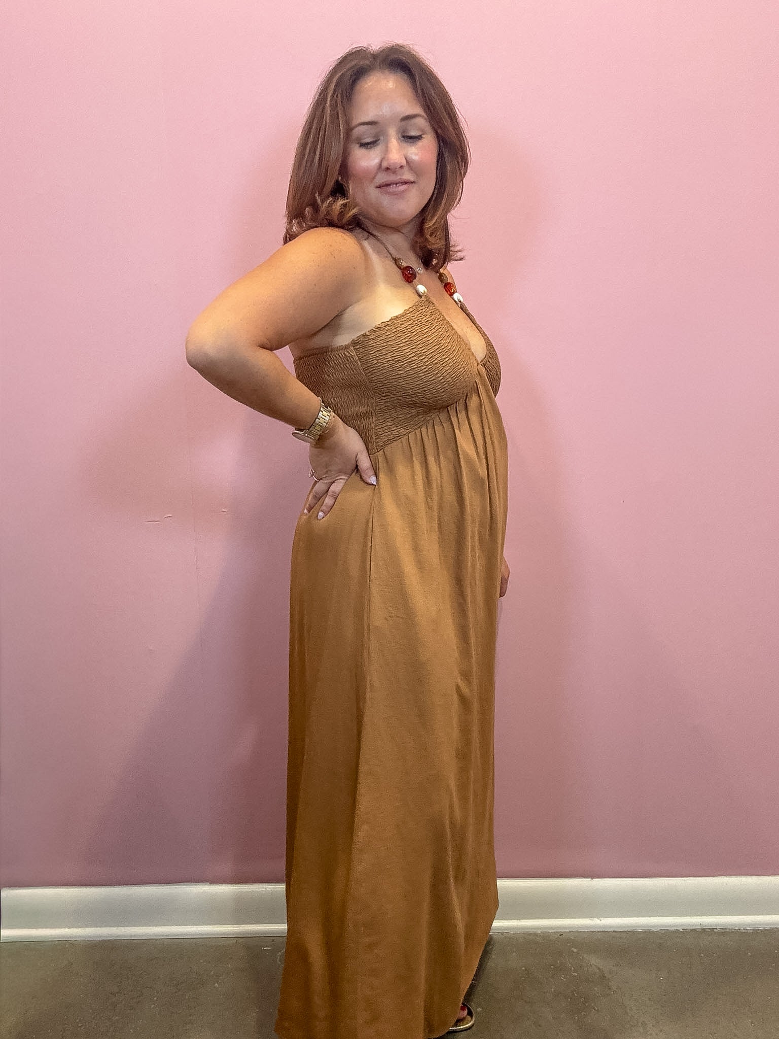 A woman modeling a sleeveless, smocked-bust maxi dress in a warm camel brown color, standing against a solid pink wall.