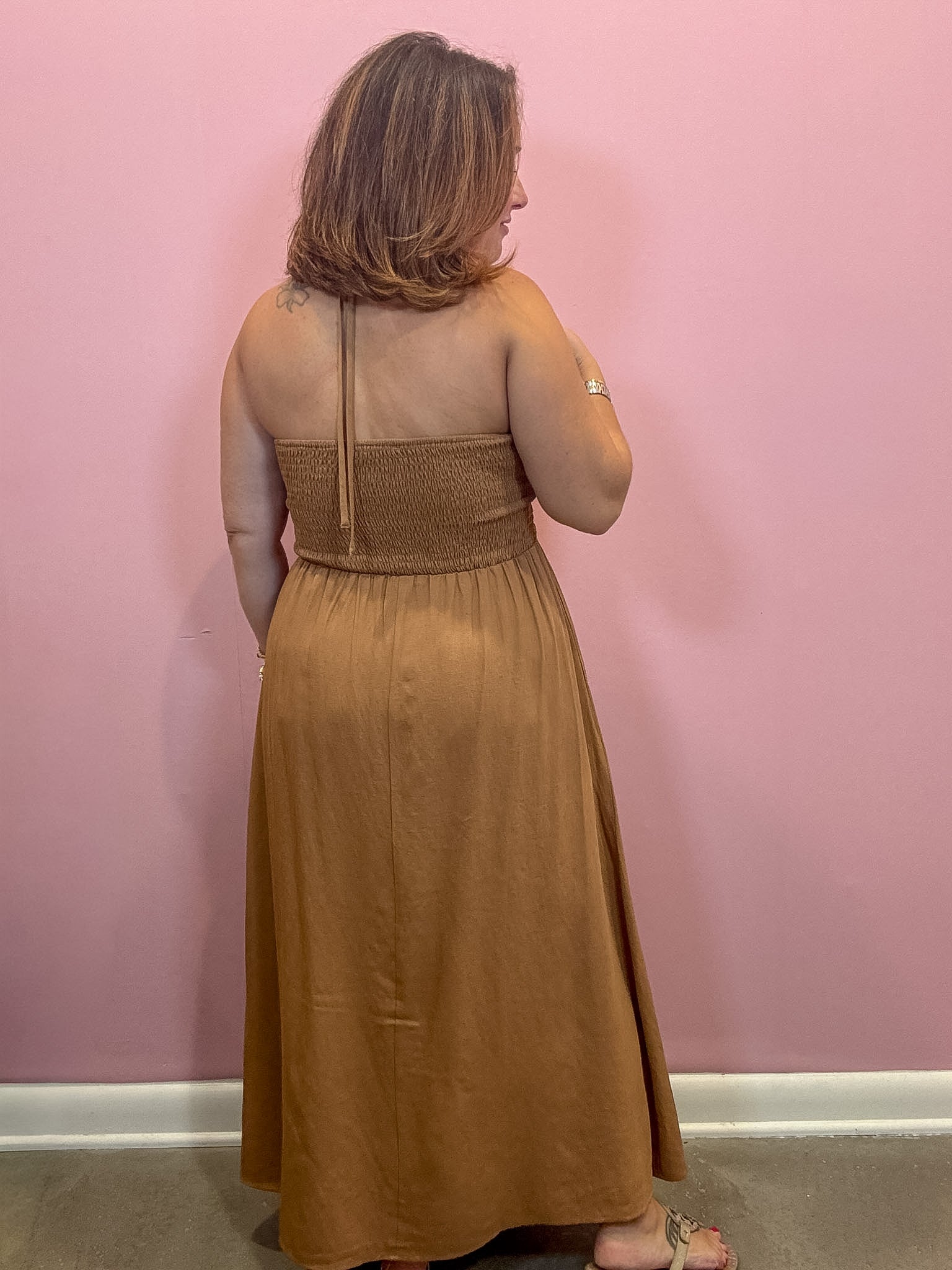 A woman viewed from behind wearing a sleeveless, smocked-bust maxi dress in a warm camel brown color, standing against a solid pink wall.