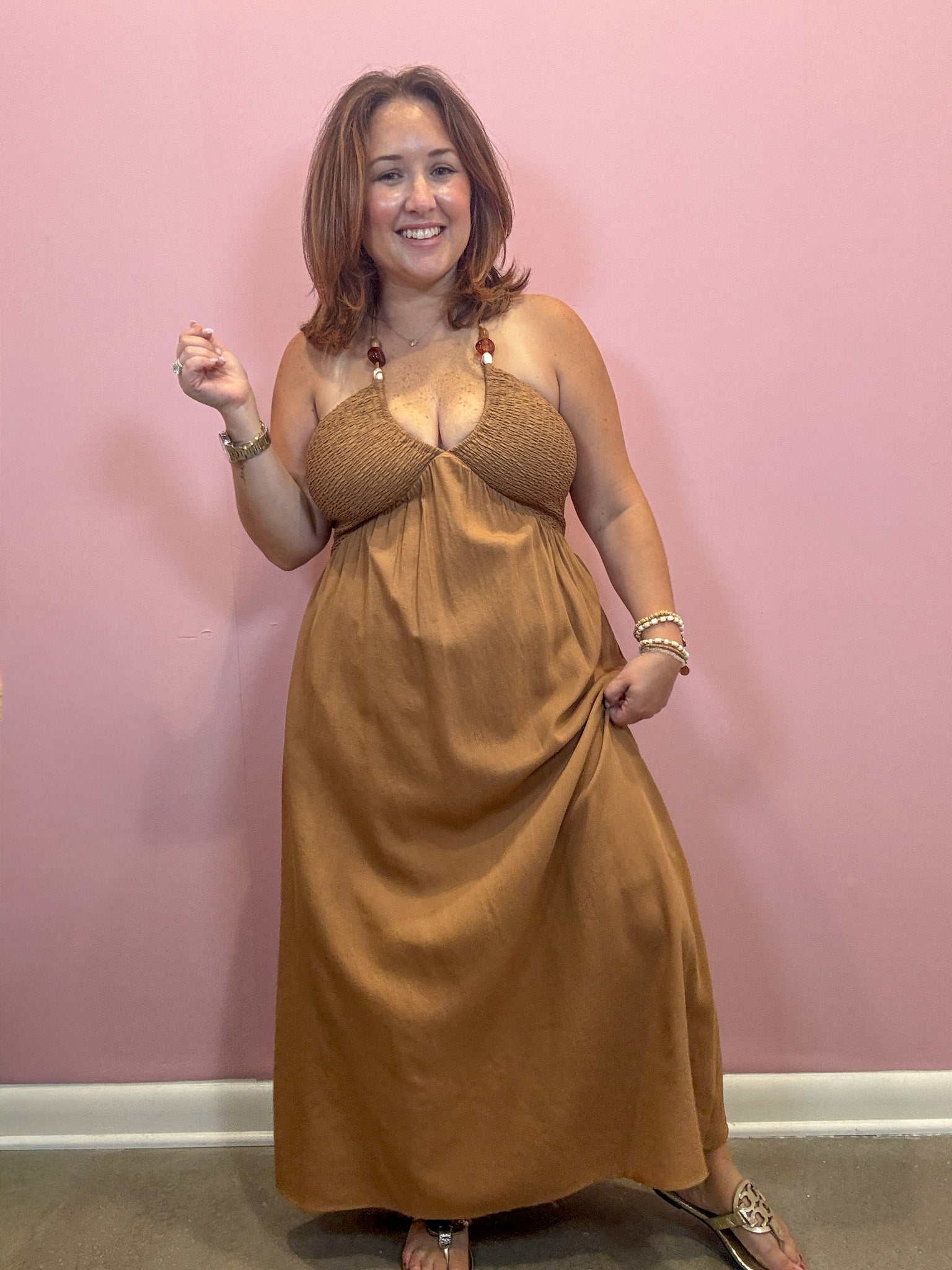 A woman modeling a brown halter-neck maxi dress with a textured bralette-style top, standing against a solid pink wall.