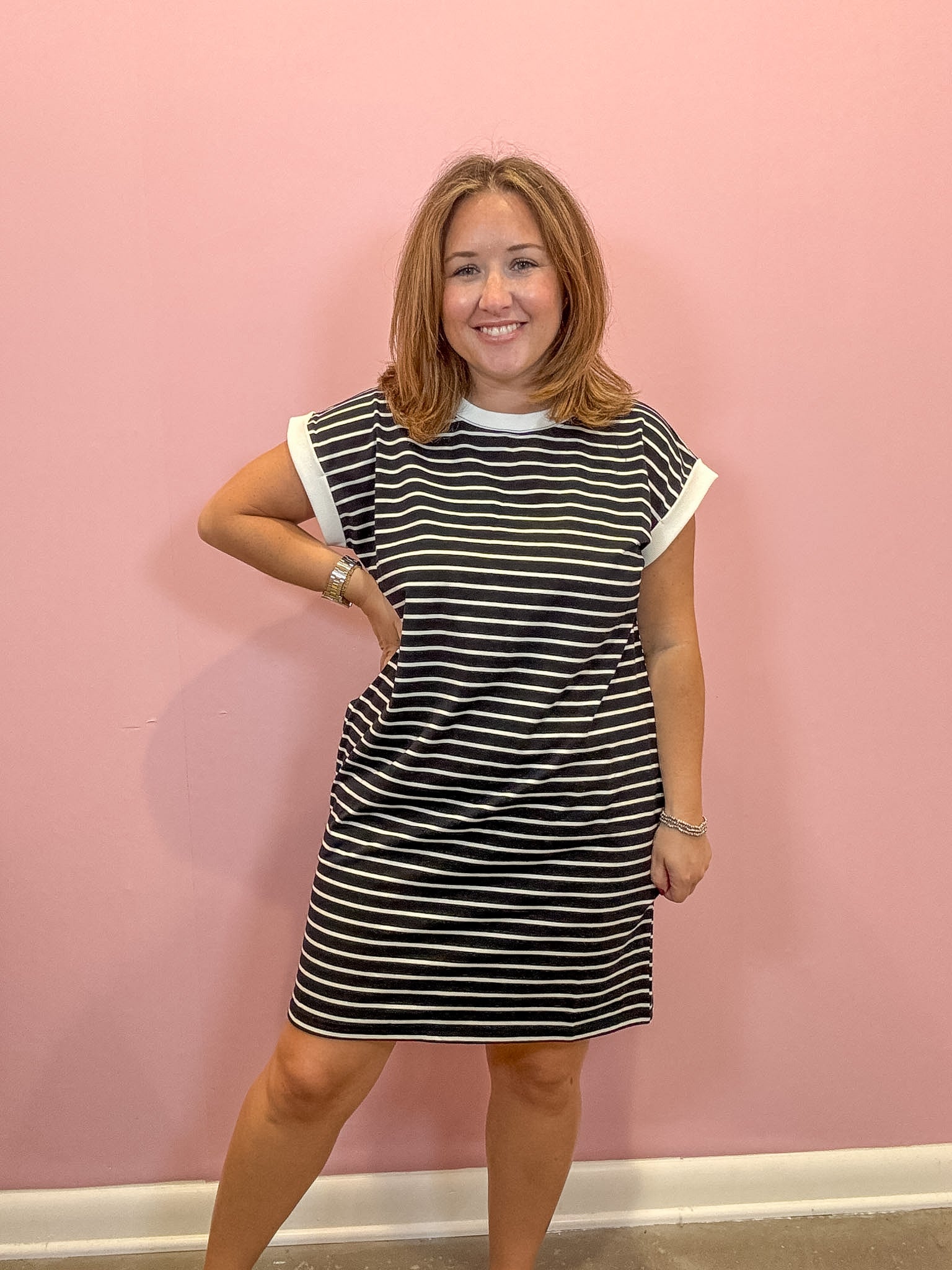 A woman modeling a short-sleeve black and white striped dress with white trim on the collar and sleeve cuffs, standing against a solid pink wall.