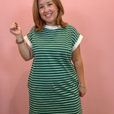 A woman modeling a green and white horizontally striped short-sleeve t-shirt dress against a solid pink wall.